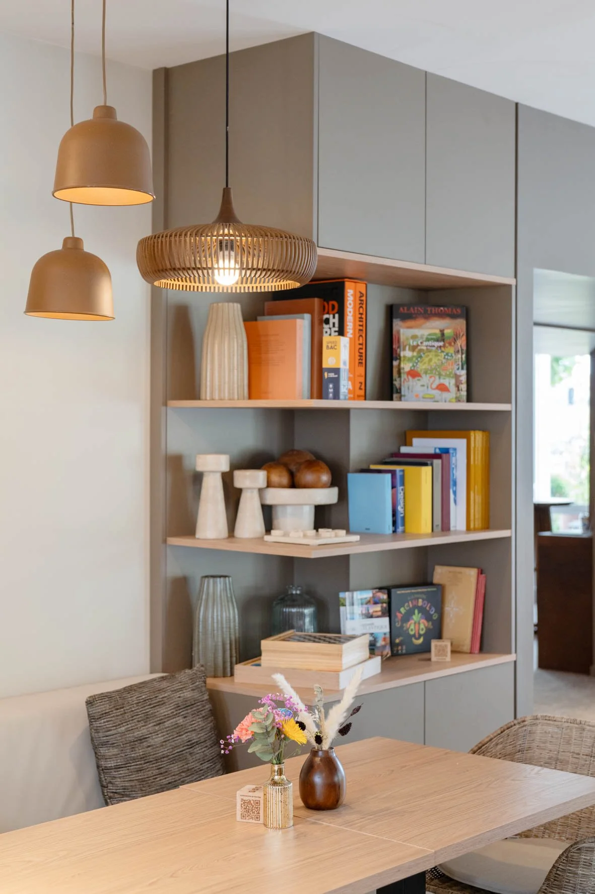 Une bibliothèque en bois gris clair avec des étagères contenant des livres de différentes couleurs et des décorations en céramique, suspendue sous un plafond à plusieurs lampes décoratives en forme de cloche en beige et en bois.