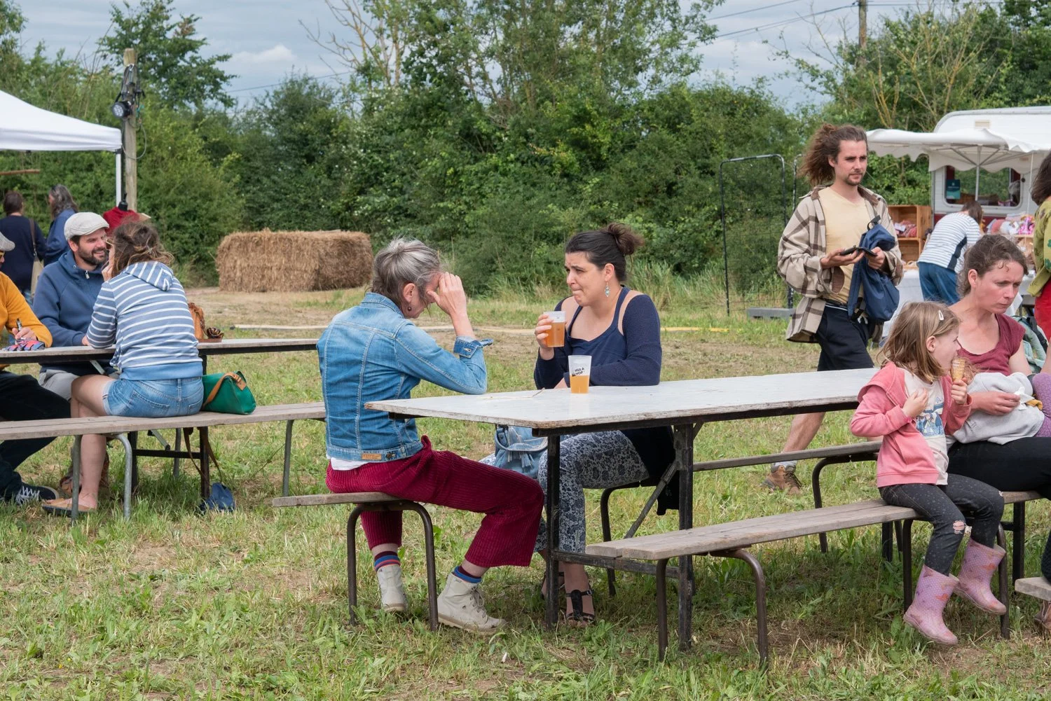 Groupe de personnes assises à une table en plein air lors d'un événement en plein air, avec des arbres en arrière-plan. Certaines personnes parlent, d'autres mangent ou boivent.