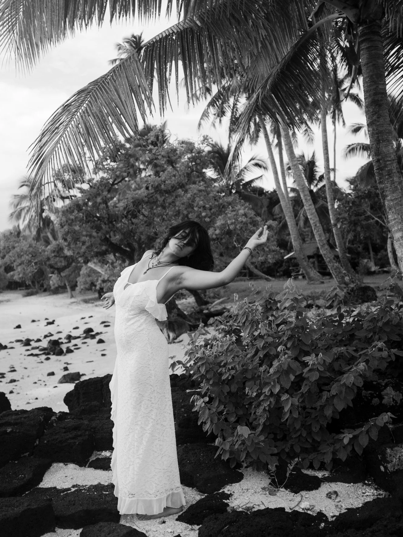 Une femme en robe longue blanche posant sur une plage avec des arbres tropicaux en arrière-plan, en noir et blanc.