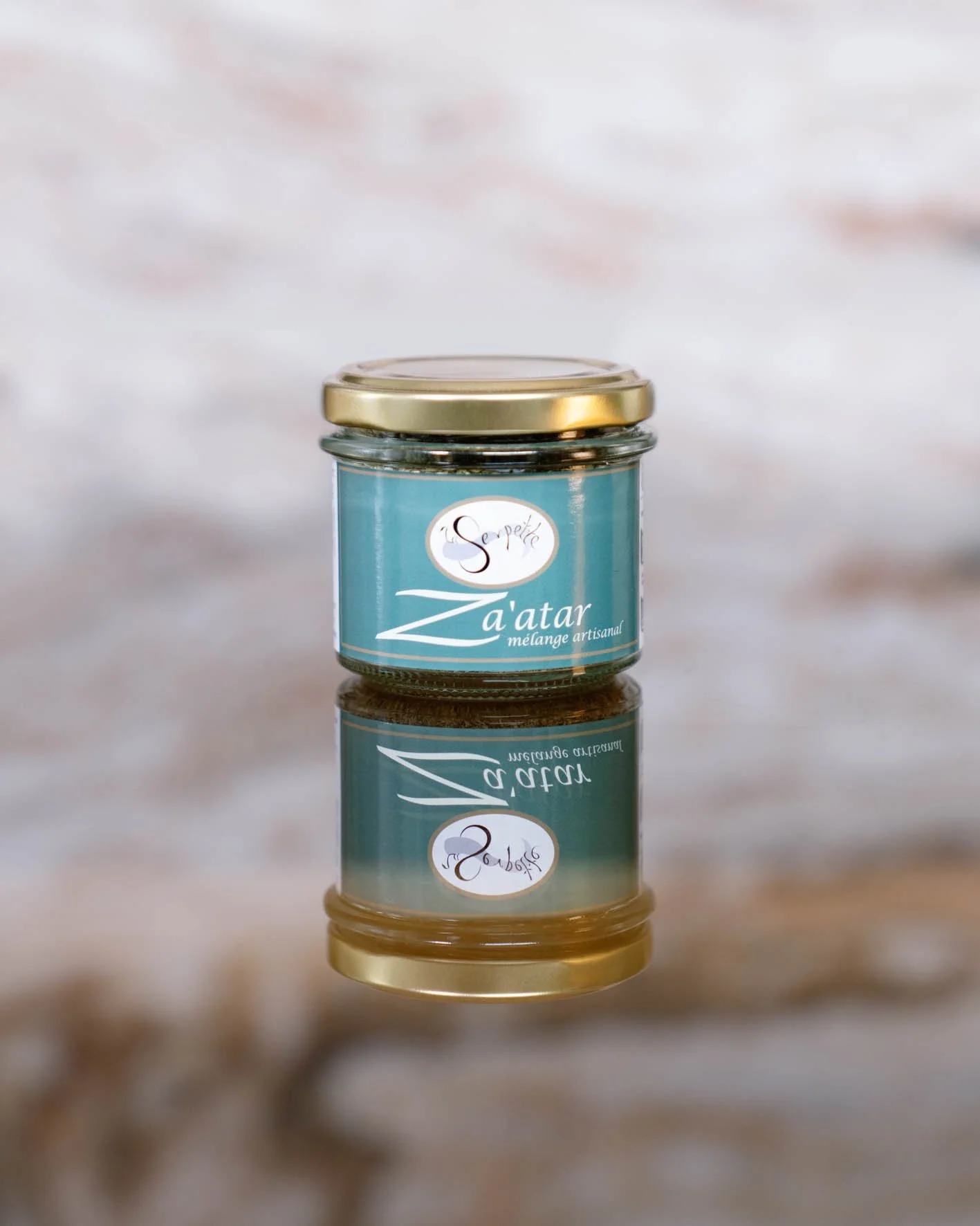 Petit pot en verre avec une étiquette bleue, contenant une pâte ou une crème, avec un couvercle or. Le pot est posé sur une surface en bois, et est reflété en dessous.