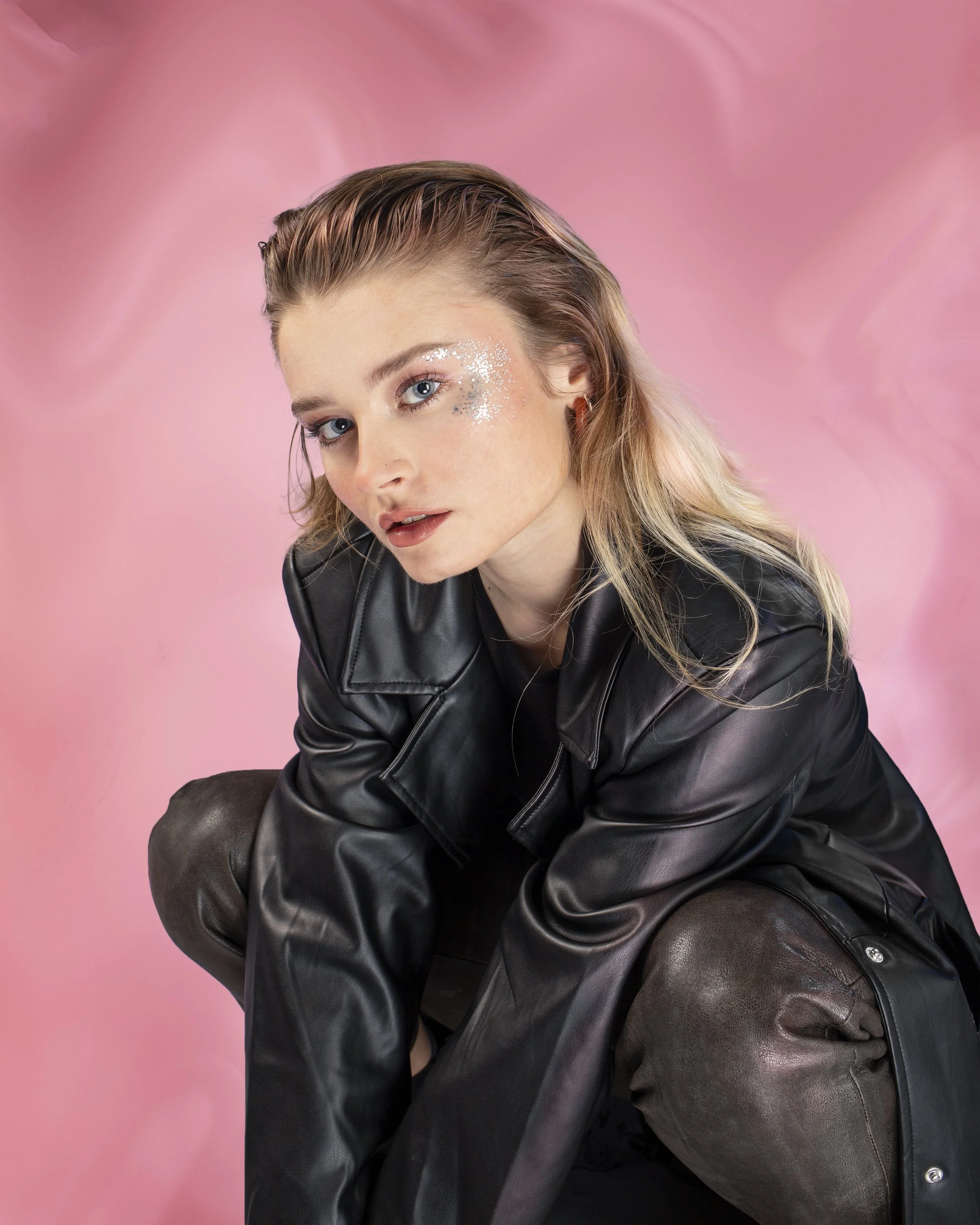 Jeune femme aux cheveux blonds, maquillage avec paillettes argentées autour de l'œil, portant un blouson en cuir noir, assise contre un fond rose vif. Photographie de mode