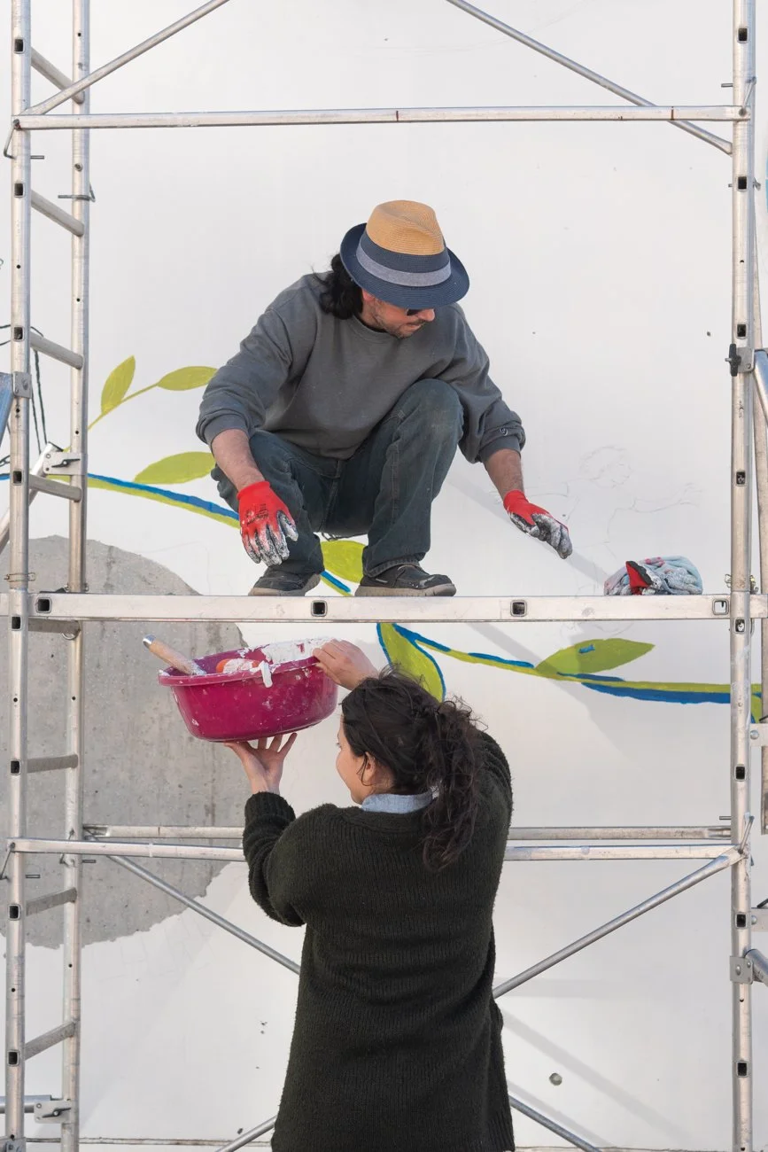 Deux personnes, un homme sur une échelle travaillant sur un mur peint avec une branche et des feuilles, et une femme passant un bol de peinture à l'homme, tous deux portant des gants.