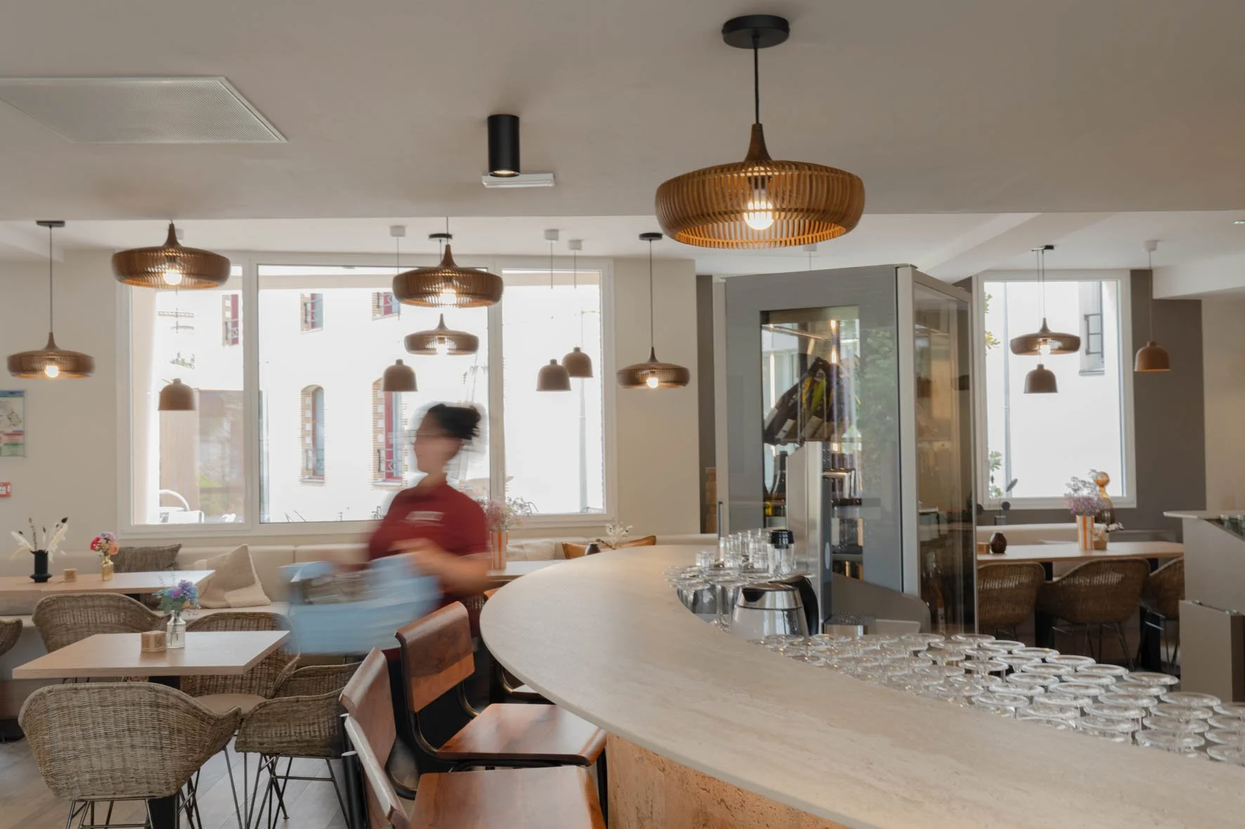 Intérieur d'un café ou restaurant lumineux avec plusieurs lampes suspendues et une femme floue en mouvement, assise à une table.