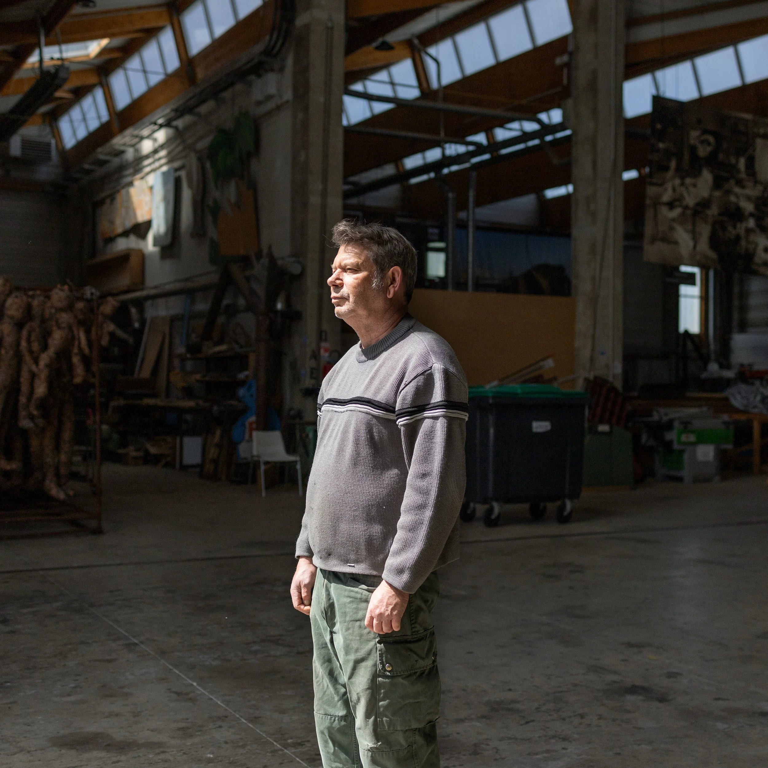 Un homme d'âge moyen porte un pull gris à rayures et un pantalon vert dans un entrepôt ou un atelier avec des poutres en bois et des matériaux stockés.