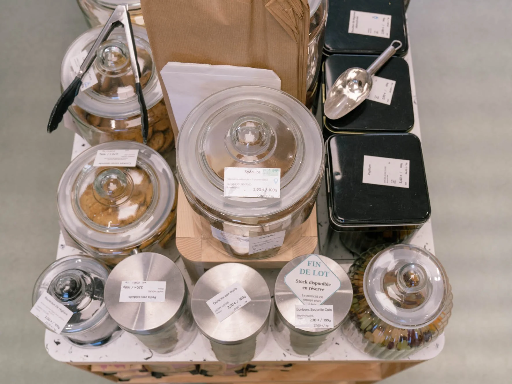 Varie contenitori di spezie e biscotti in barattoli di vetro con etichette, alcuni barattoli con coperchi metallici rotondi, alcuni con etichette di prezzo e informazioni in francese, disposti su un tavolo di commercio o negozio.