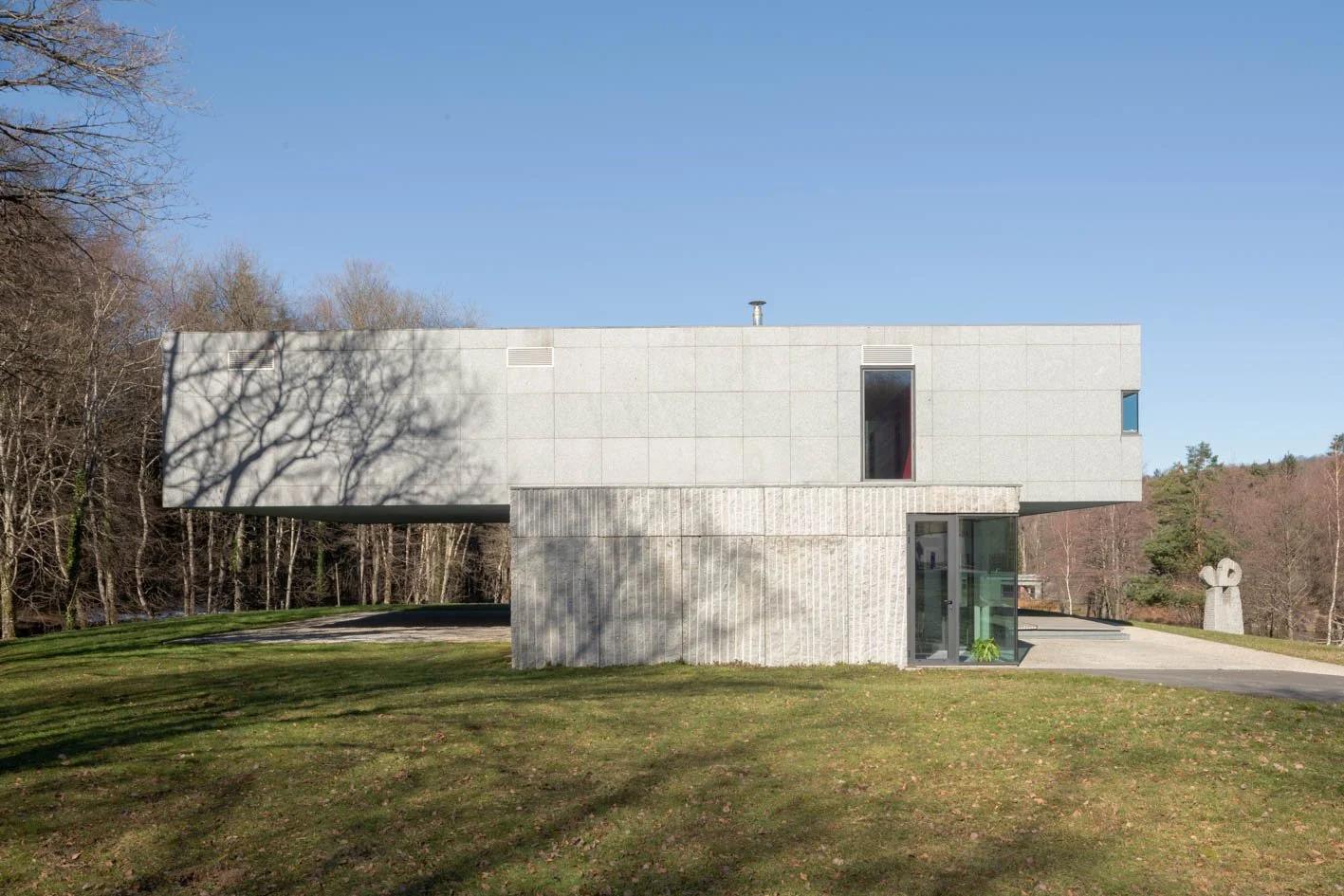 Une maison moderne avec une façade en pierre et en verre, située dans un espace vert avec des arbres en arrière-plan, un ciel clair et ensoleillé.