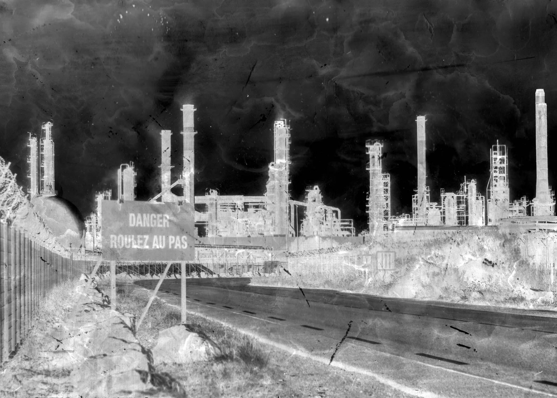 Une usine industrielle avec plusieurs cheminées, des structures métalliques et un panneau de signalisation en français indiquant « Danger, ROULEZ AU PAS ». Une photo en noir et blanc détériorée à but artistique.