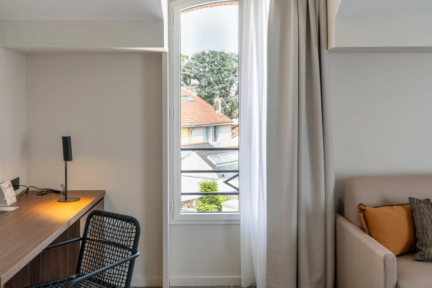 Une pièce lumineuse avec une fenêtre donnant sur des toits et des arbres, un canapé beige avec des coussins, et un bureau en bois avec une lampe