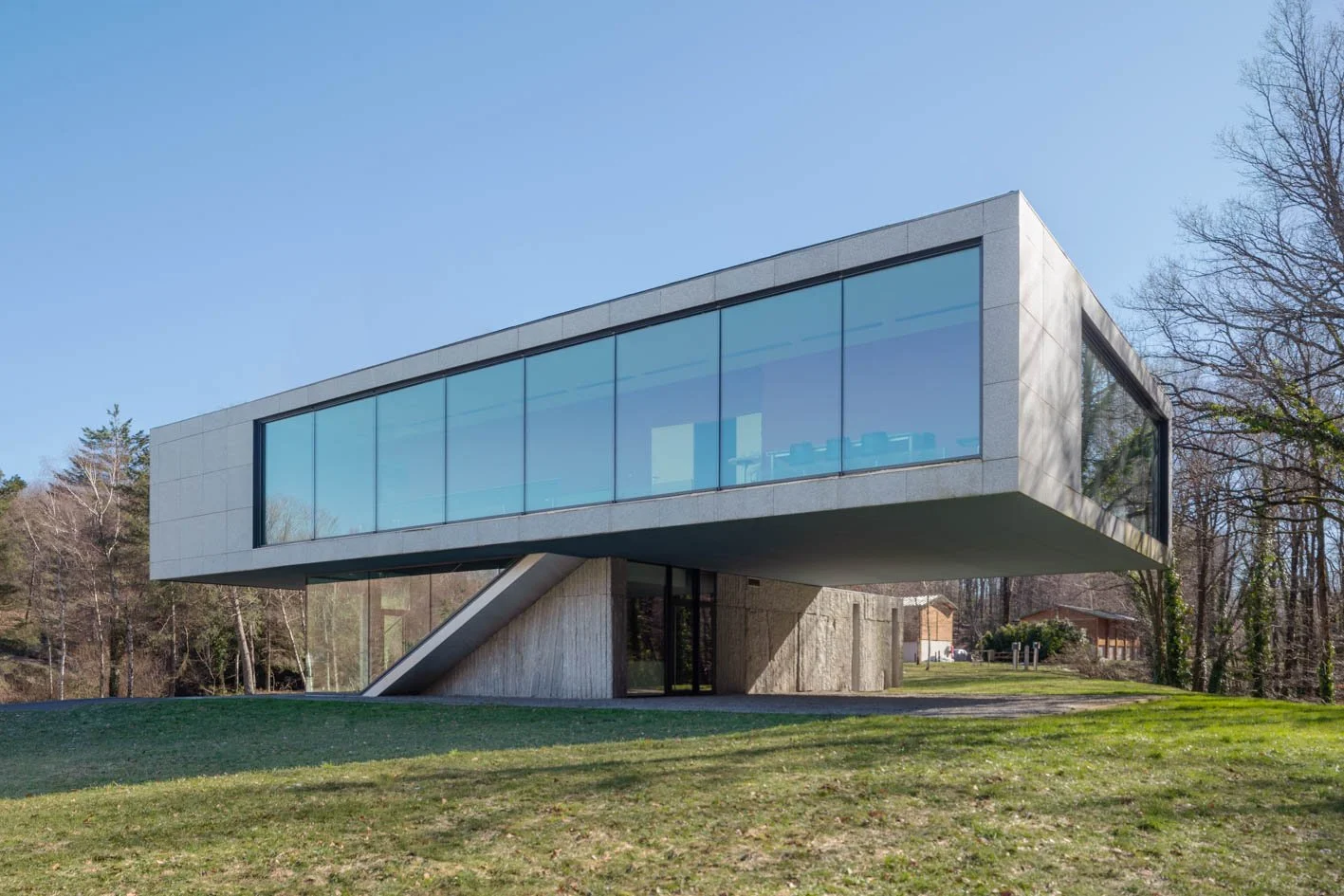 Une maison moderne en béton et verre, construite sur pilotis, située dans un environnement boisé avec un ciel bleu clair.
