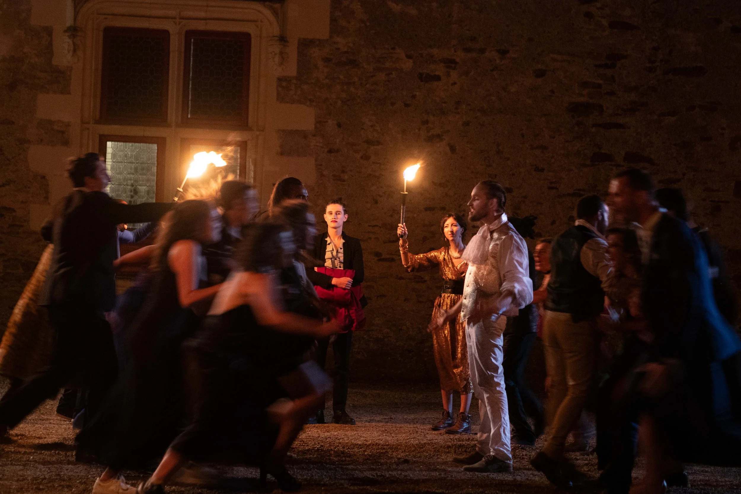 Groupe de personnes lors d'une fête en soirée, certaines avec des torches allumées, dans une cour avec un mur en pierre et une grande fenêtre en arrière-plan.