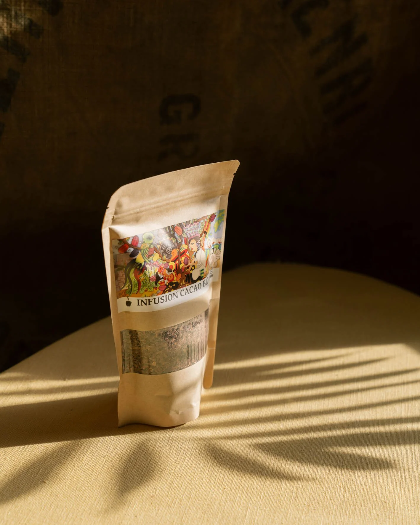 Sachet de pâte à infusion cacao bio placé sur une surface en tissu beige avec des rayons de lumière qui créent des ombres.
