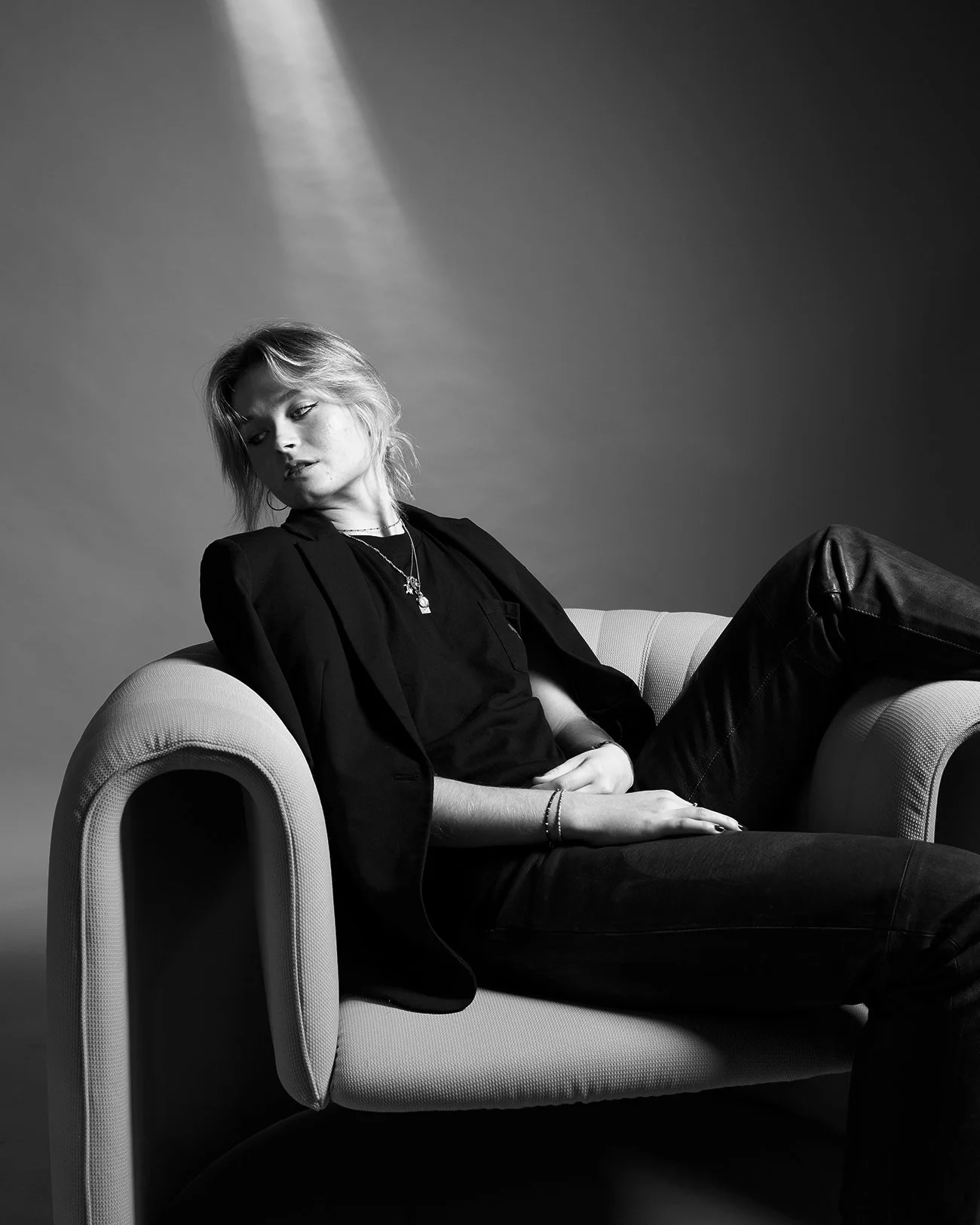 Une jeune femme avec des cheveux courts, portant une veste noire et un t-shirt noir, assise de manière décontractée sur un canapé en cuir clair, dans une ambiance sobre en noir et blanc.