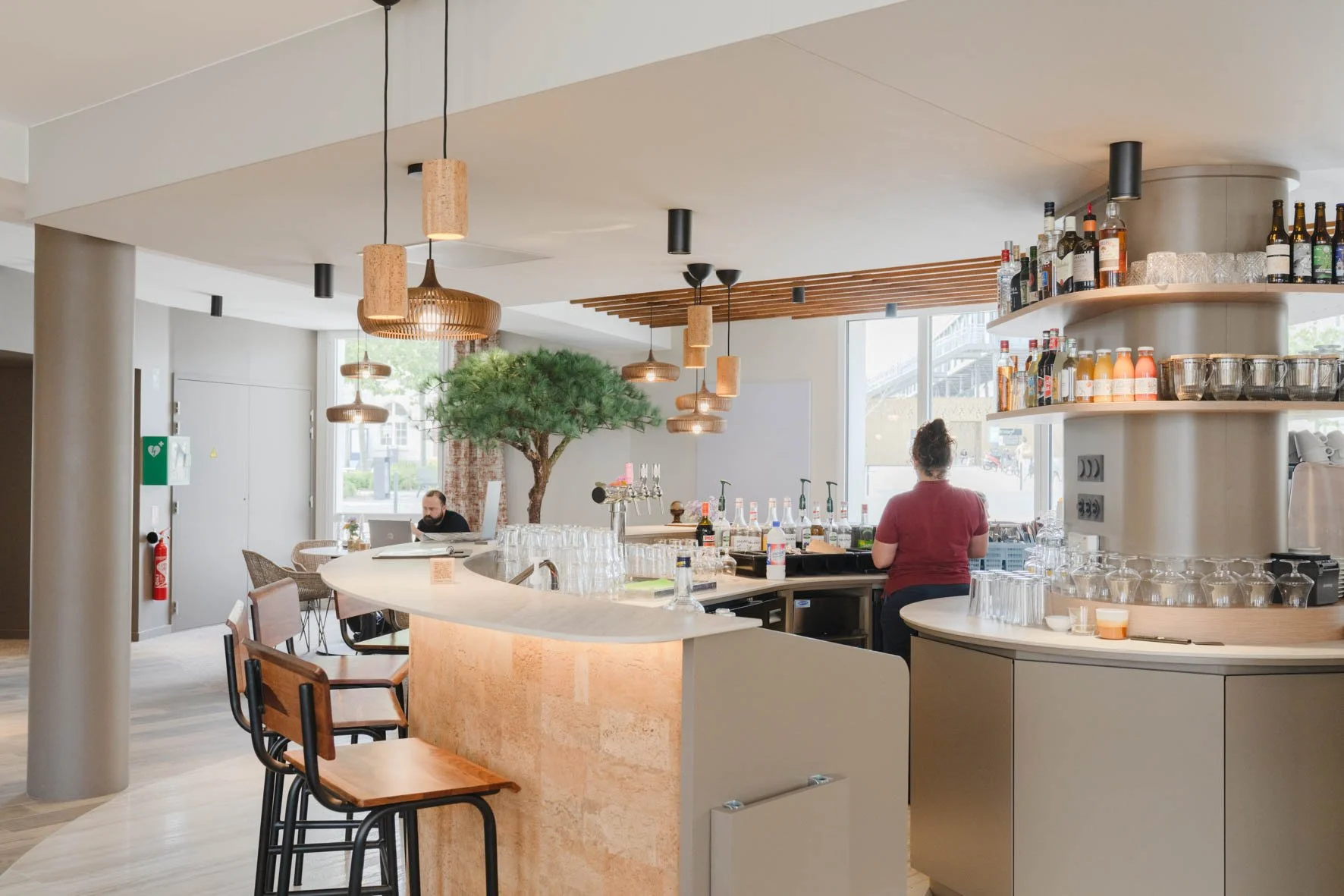 Intérieur d'un bar moderne avec un comptoir en marbre, étagères de bouteilles de boissons alcoolisées, et deux personnes derrière le bar, une assise à une table et une autre en train de préparer des boissons, décoration végétale, éclairage suspendu e