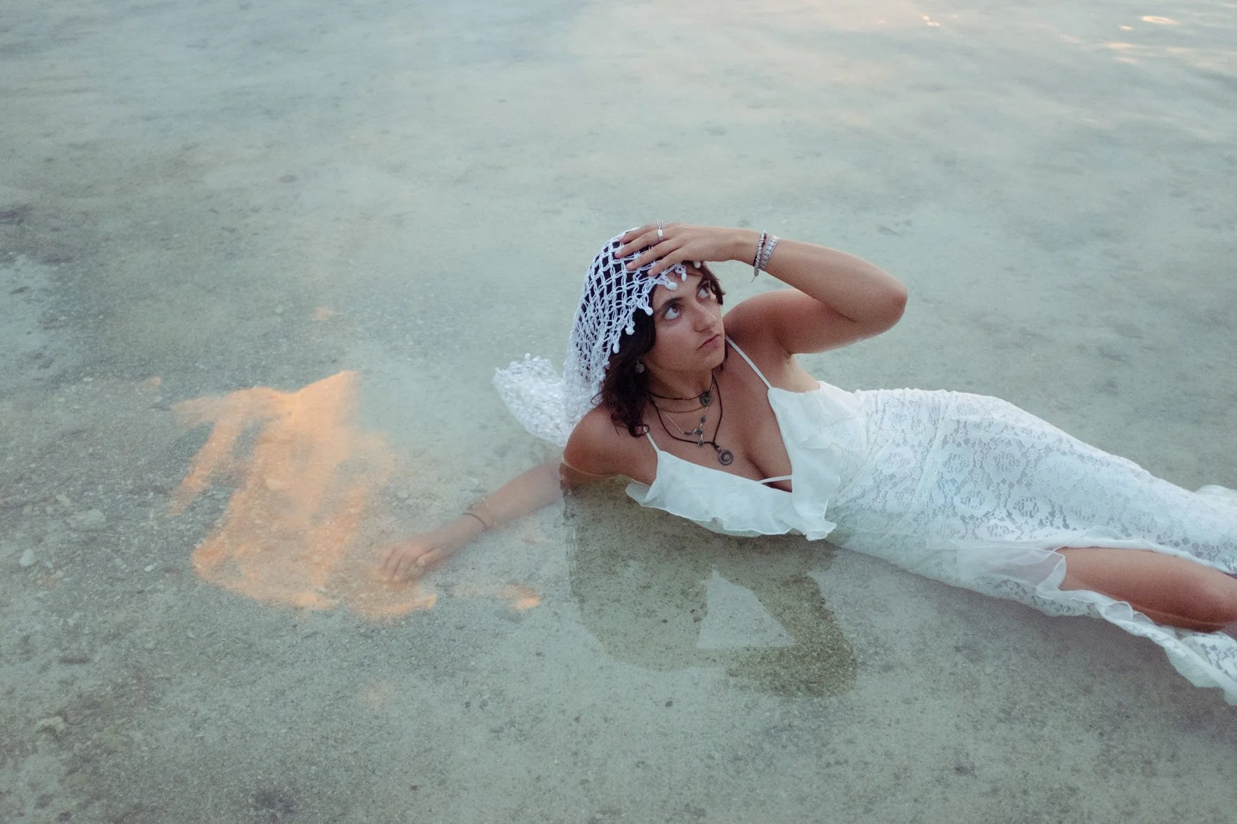 Femme en robe blanche allongée dans l'eau de la mer ou d'un lac, portant un voile blanc, avec des bijoux, regardant vers le haut.