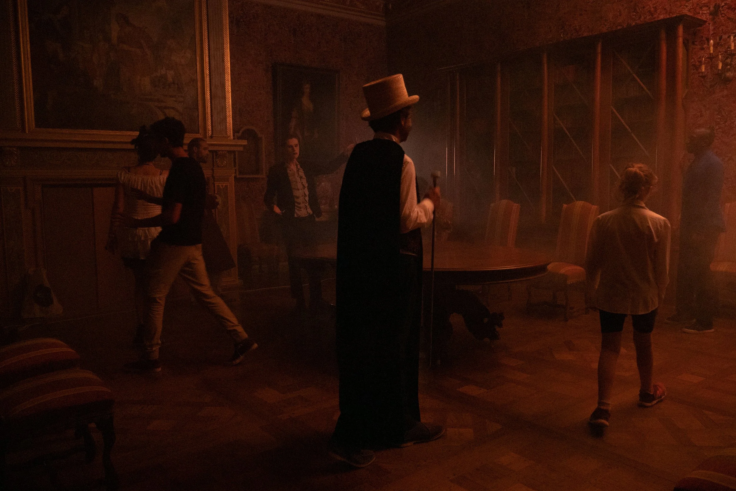 Une scène sombre dans une pièce richement décorée avec plusieurs personnes, dont une portant un haut-de-forme et un costume, évoquant une ambiance de spectacle ou de performance historique.