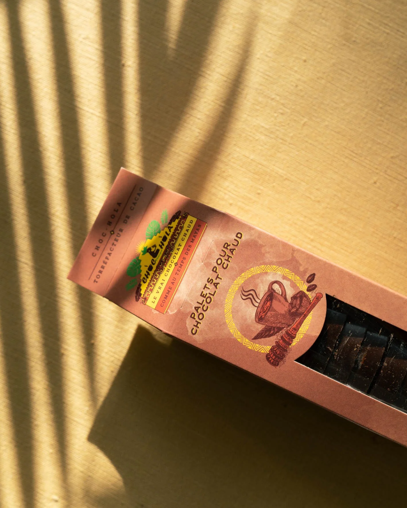 Boîte de chocolat chaud de la marque valrhona, couleur rose, avec illustration de cacao et de tasse de chocolat chaud, posé sur une surface beige.