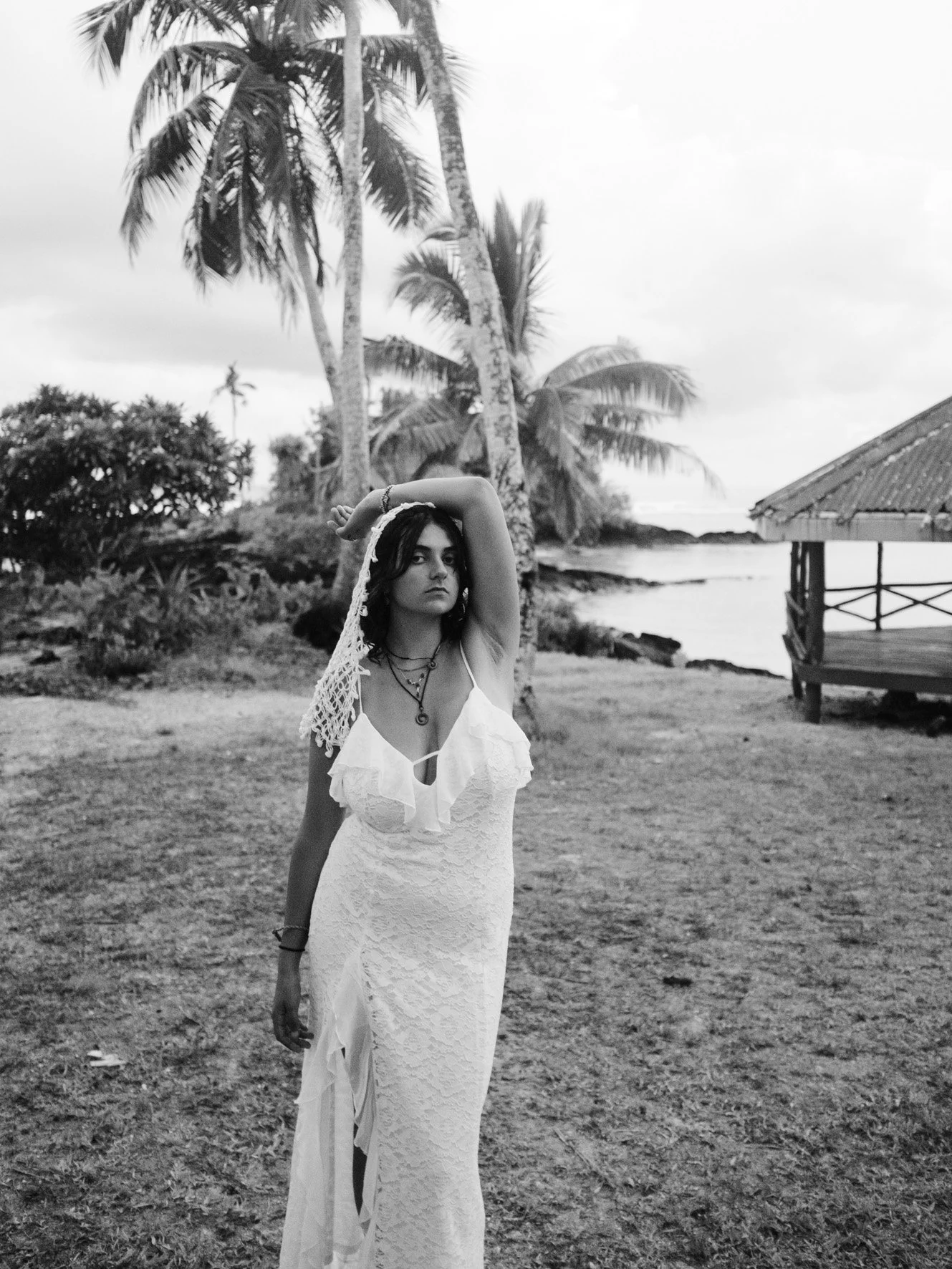 Une jeune femme en robe blanche et avec un chapeau en crochet se tient devant des palmiers sur une plage, dans un environnement tropical
