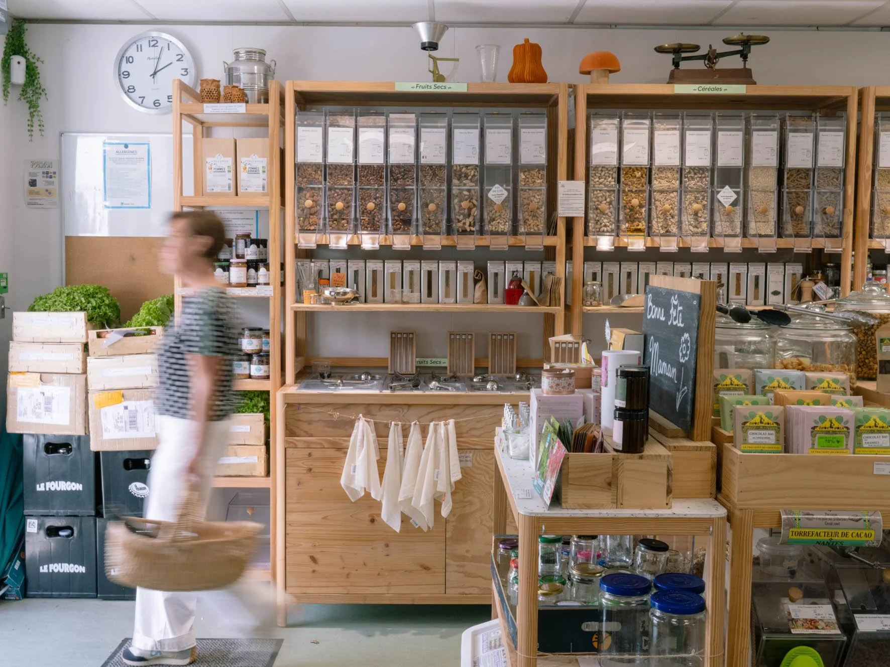 Rayons de magasin bio avec des bocaux de fruits secs, céréales et chocolats, et un client portant un panier de courses en marche.