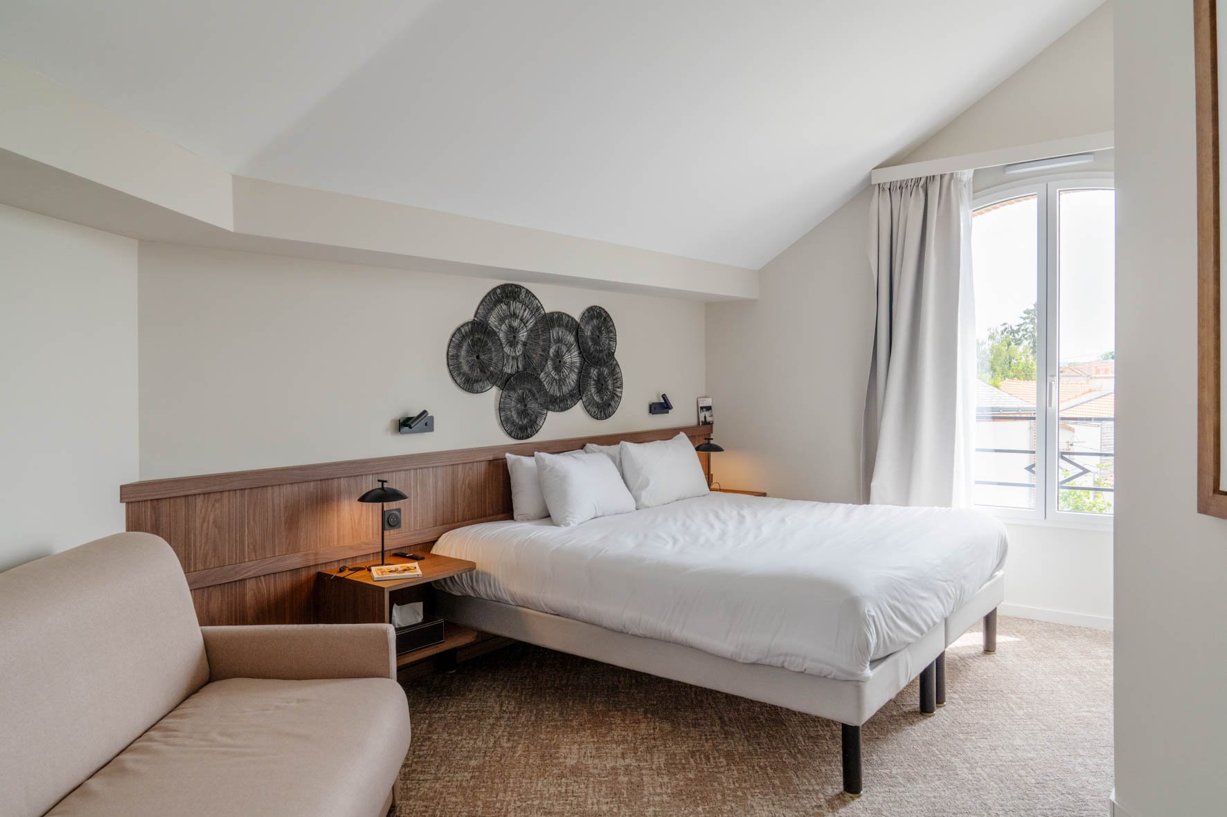 Chambre d'hôtel lumineuse avec un lit blanc, un fauteuil beige, un mur avec décoration en métal noir, une fenêtre avec rideaux blancs, vue sur des toits et des arbres.