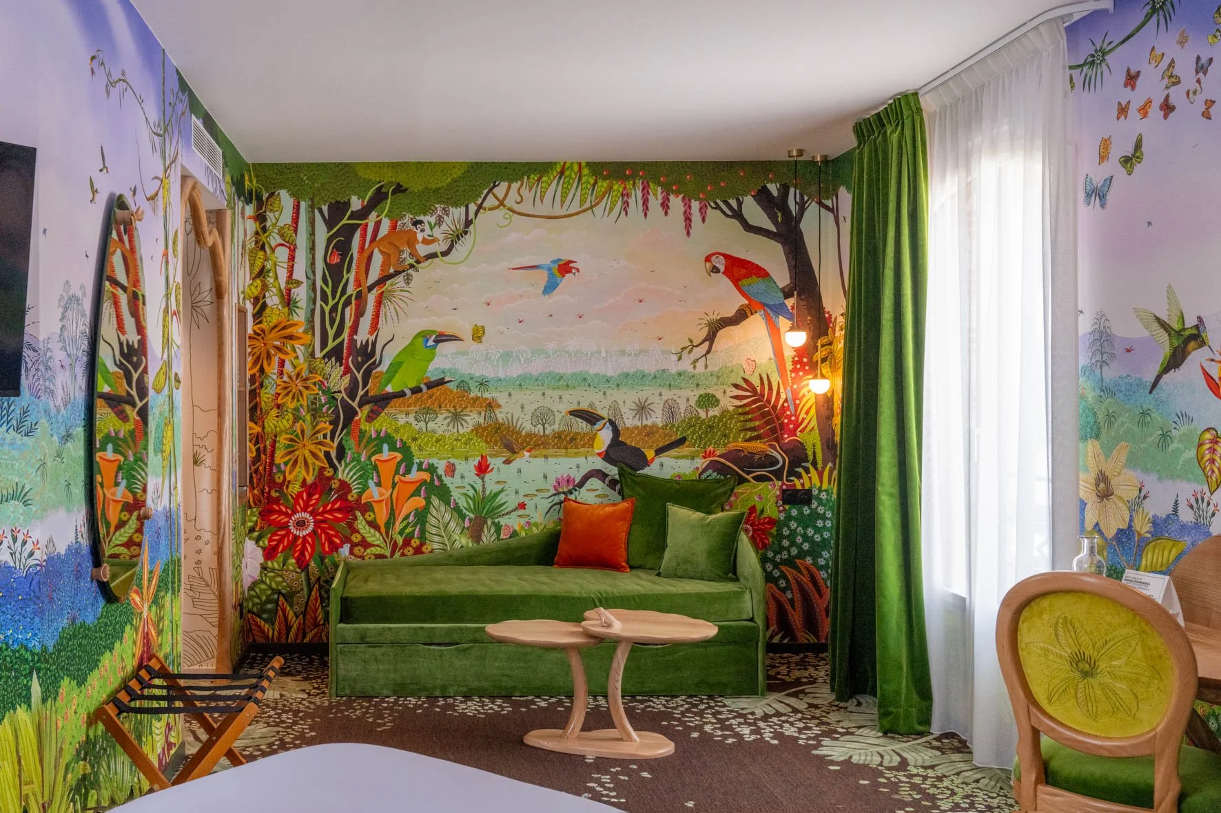 Une chambre décorée avec une peinture murale représentant une forêt tropicale avec des oiseaux et des singes, comprenant un canapé vert, des coussins orange et vert, et une fenêtre avec des rideaux verts et blancs.