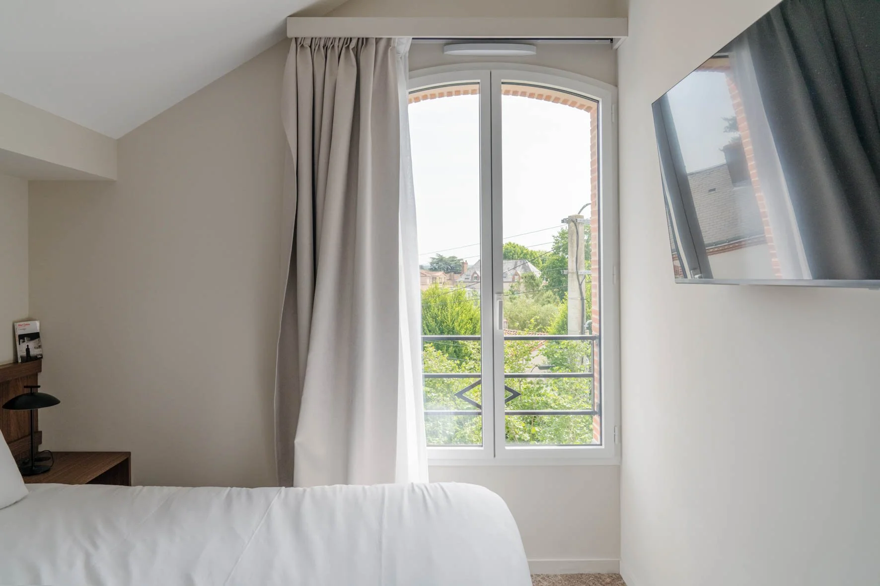 Une vue d'une chambre avec une grande fenêtre laissant entrer beaucoup de lumière naturelle. Il y a des rideaux blancs semi-ouvert et une télévision fixée au mur à droite.