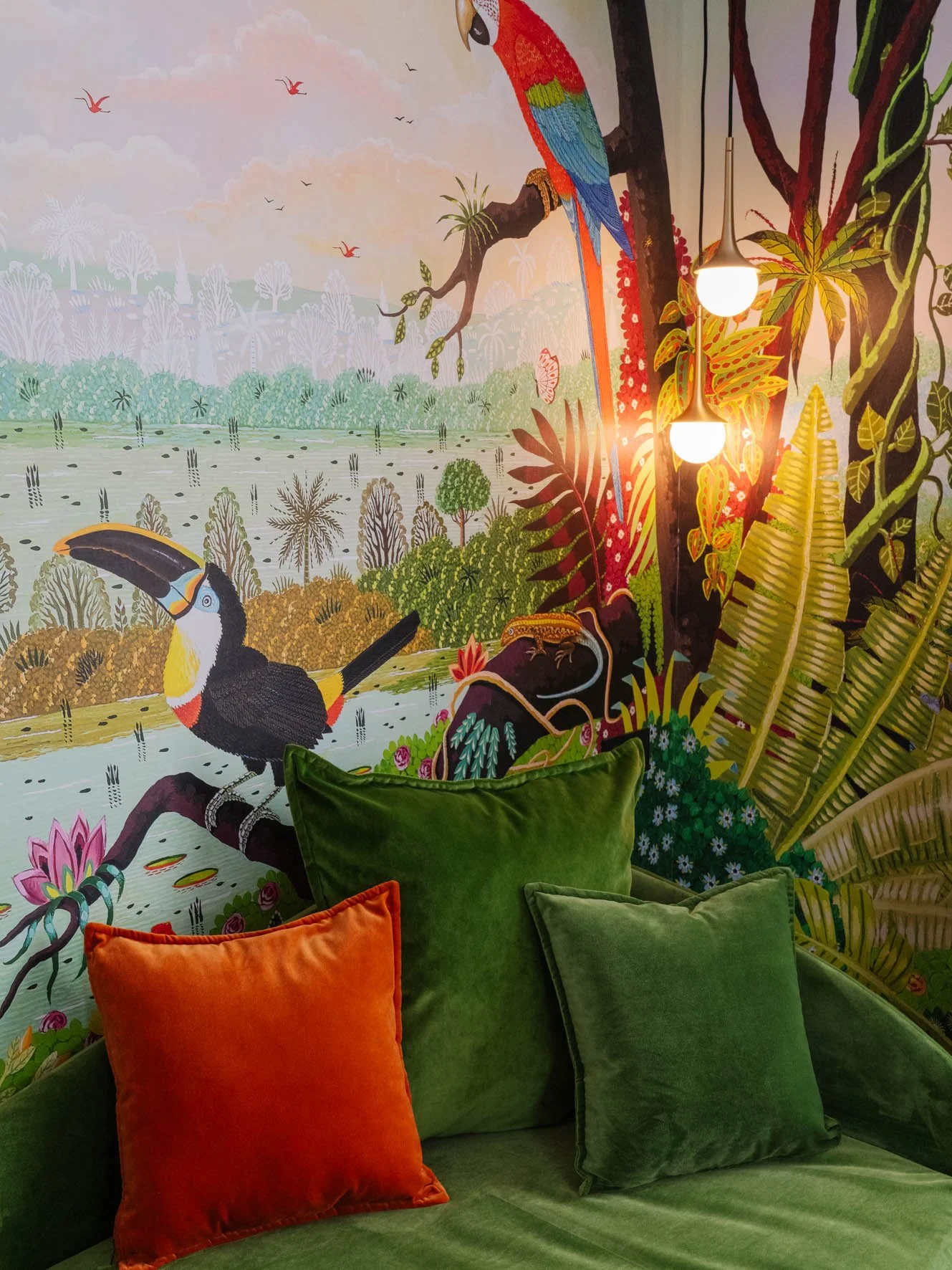 Mur peint avec une scène de jungle avec un toucan et des animaux exotiques, entouré de coussins verts et orange sur un canapé.