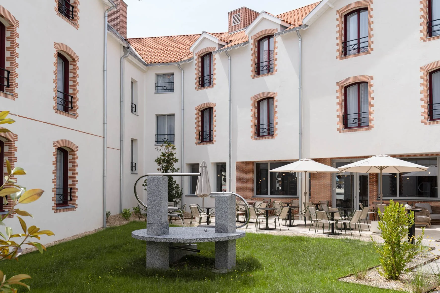 Cour intérieure d'un immeuble résidentiel avec des fenêtres, des parasols et des chaises, un jeune arbre, un puit en pierre et de la pelouse verte.