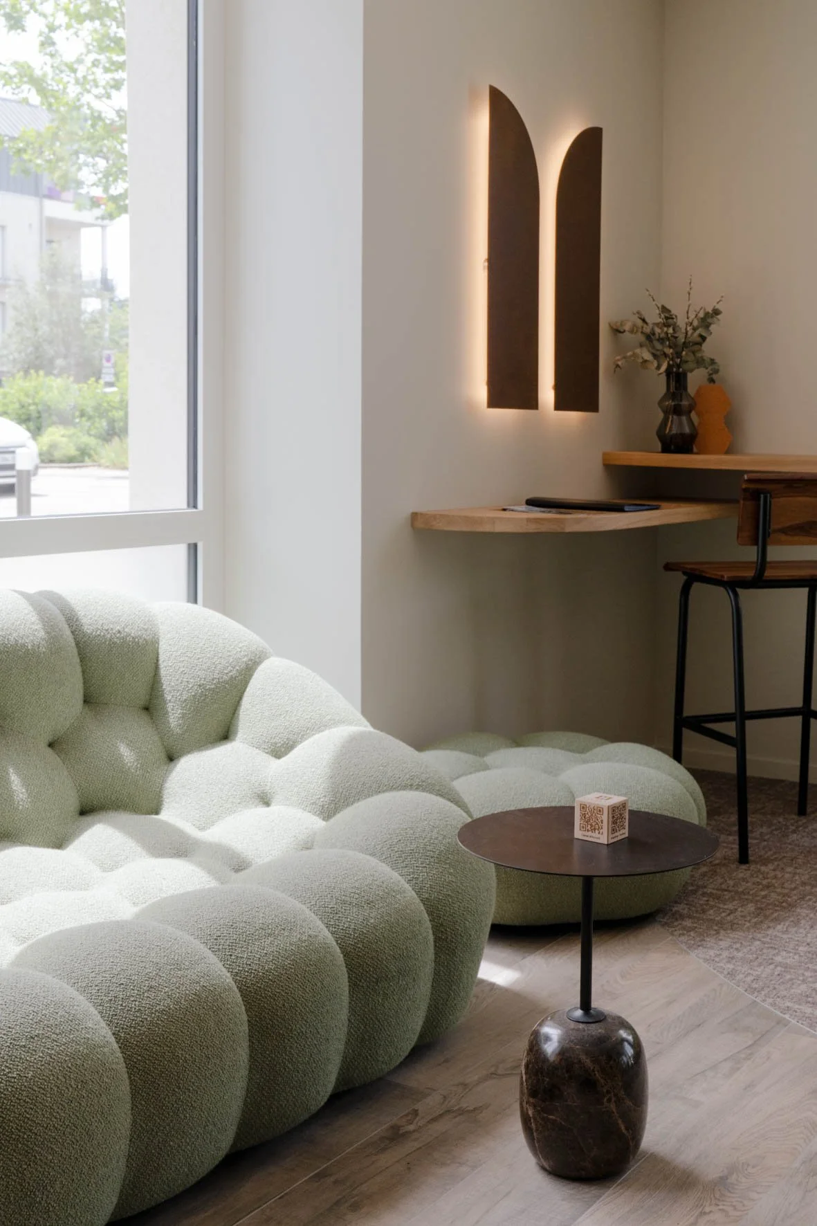 Intérieur moderne avec un canapé vert mou en forme de bulles, une petite table ronde en marbre foncé avec un cube QR code dessus, un mur blanc avec une décoration lumineuse en forme de deux arches, une table en bois avec une vase et des plantes séché