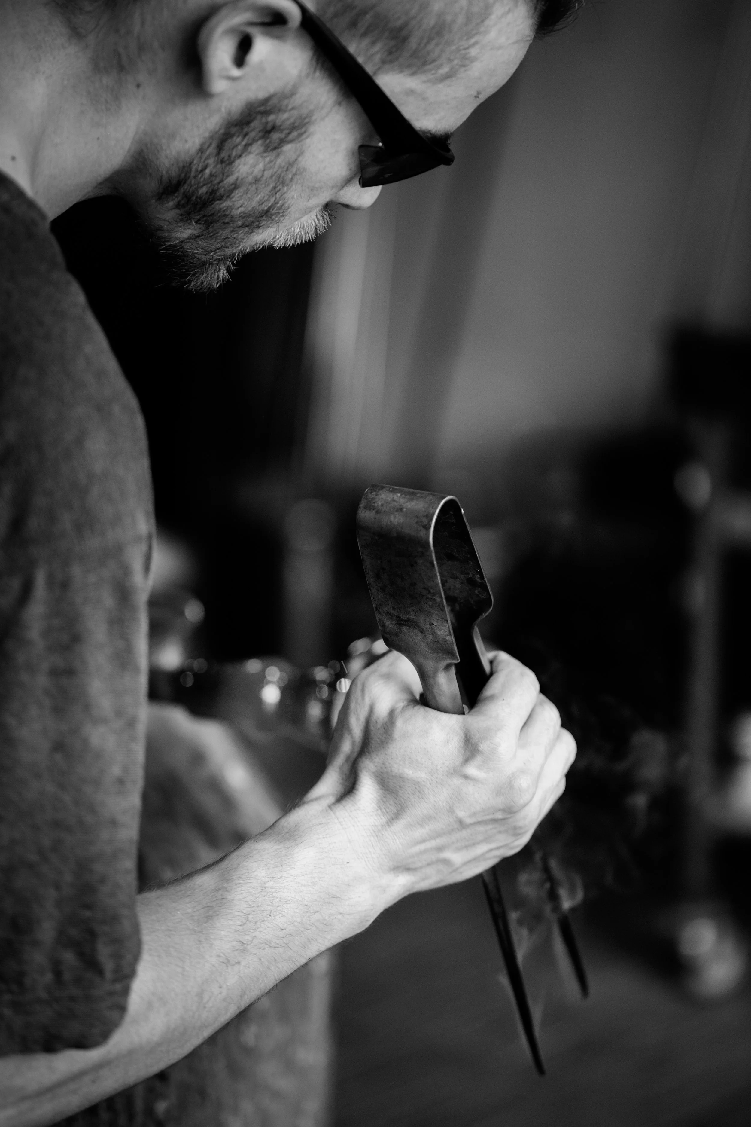 Un homme portant des lunettes tient un outil métallique en main et travaille sur une pièce métallique, dans un environnement probablement artisanal ou industriel.