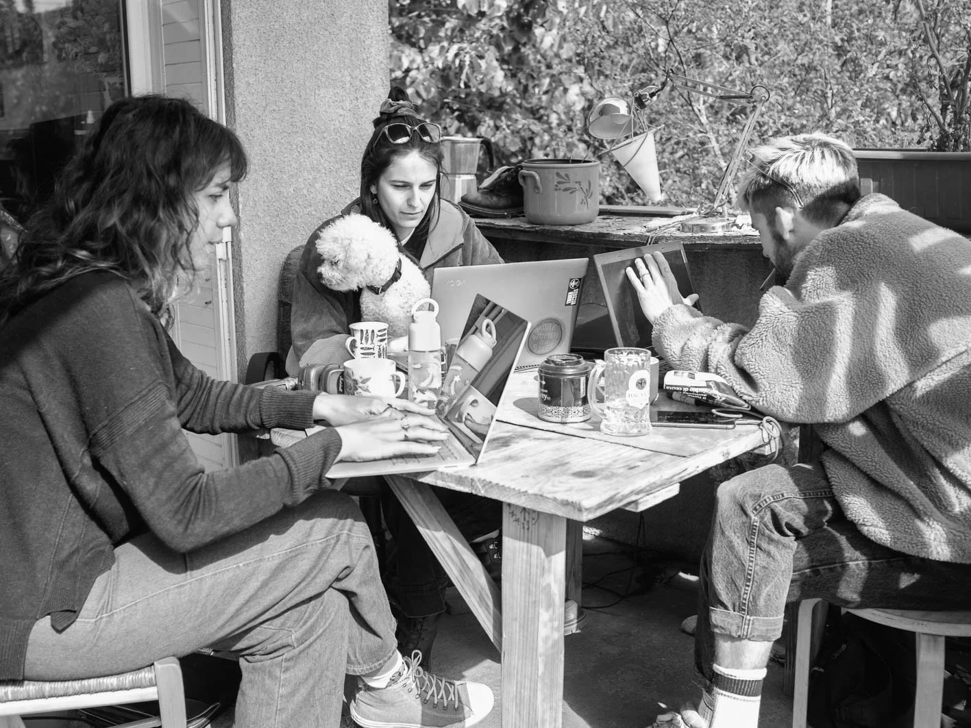 Quatre personnes et un chien assis autour d'une table en bois, utilisant des ordinateurs portables, avec des tasses et des objets divers, sur un patio extérieur.