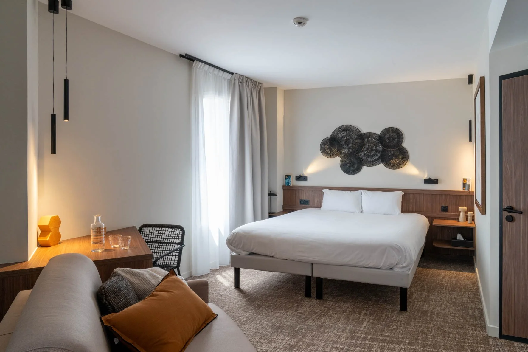Chambre d'hôtel moderne avec lit double, décoration en bois, mur d'art métallique, canapé beige avec coussins, bureau en bois avec lampe et décoratif, et grandes fenêtres avec rideaux blancs