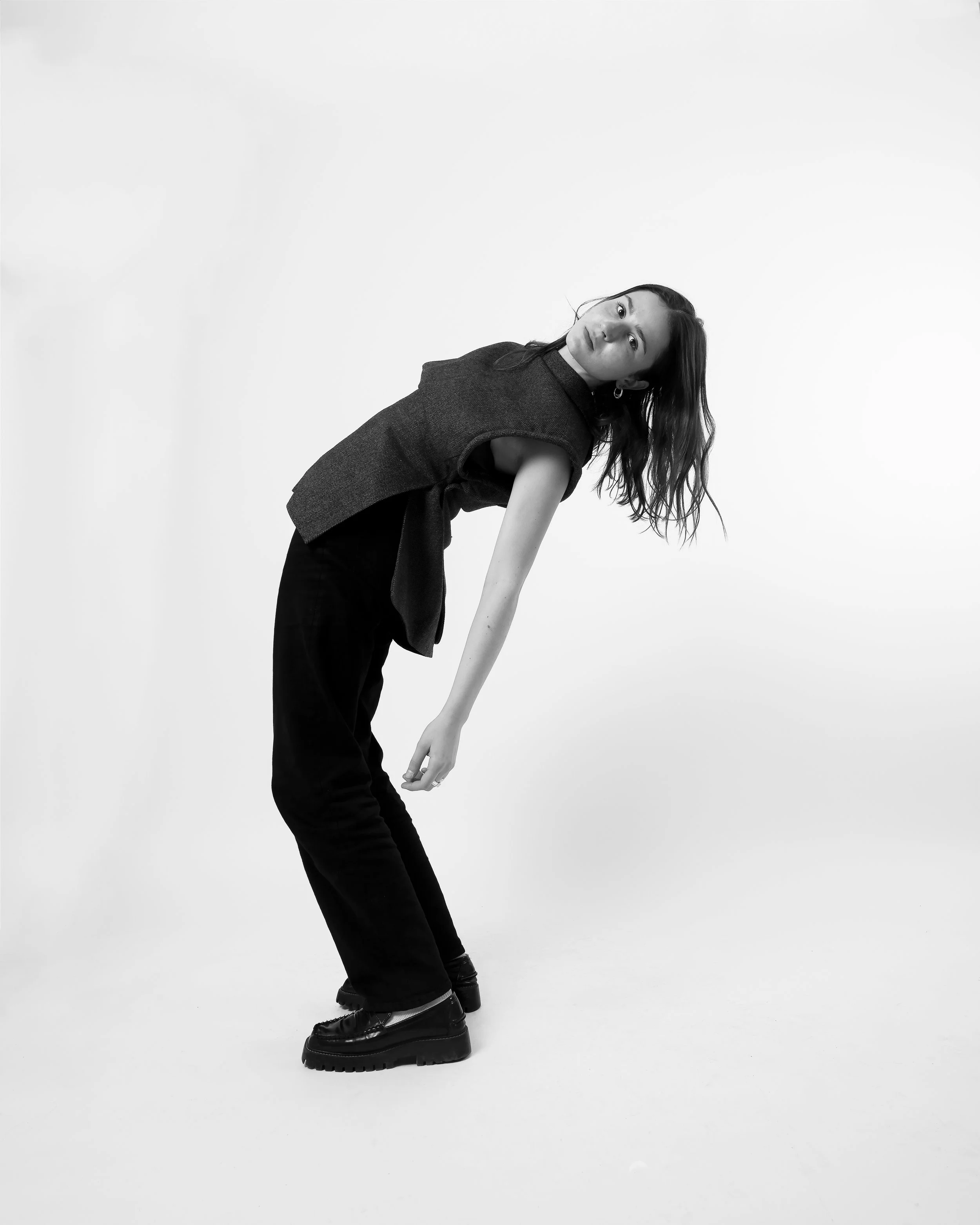 Une femme en vêtements sombres pose de biais, portant des bottes noires élégantes, sur fond blanc. Photographie de mode pour mettre en avant la vest upclycler réalisé par Joalie Chantreau