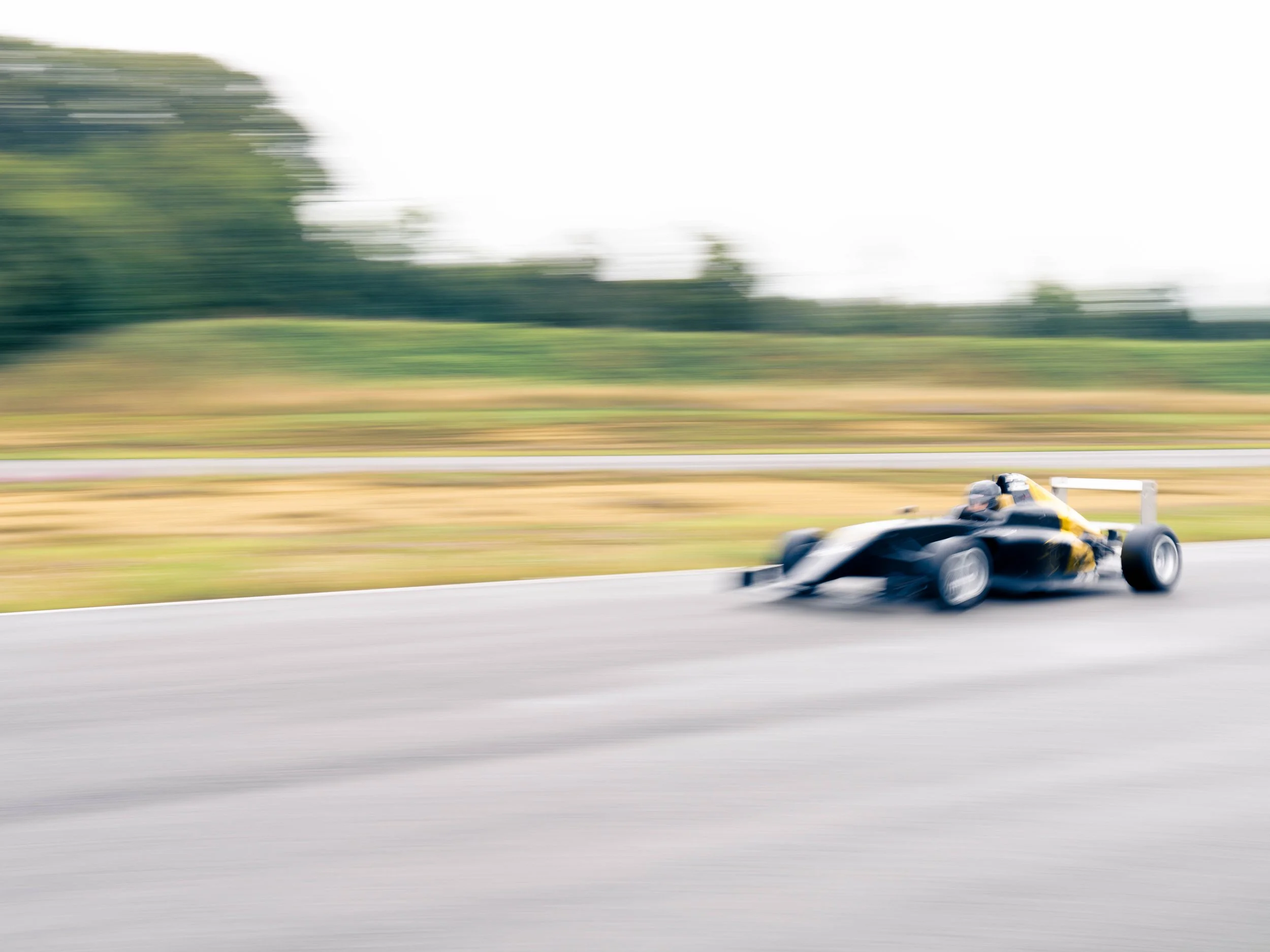 Voiture de course noire et jaune roulant rapidement sur une route, floue en raison de la vitesse, avec un fond de paysage vert et blanc.
