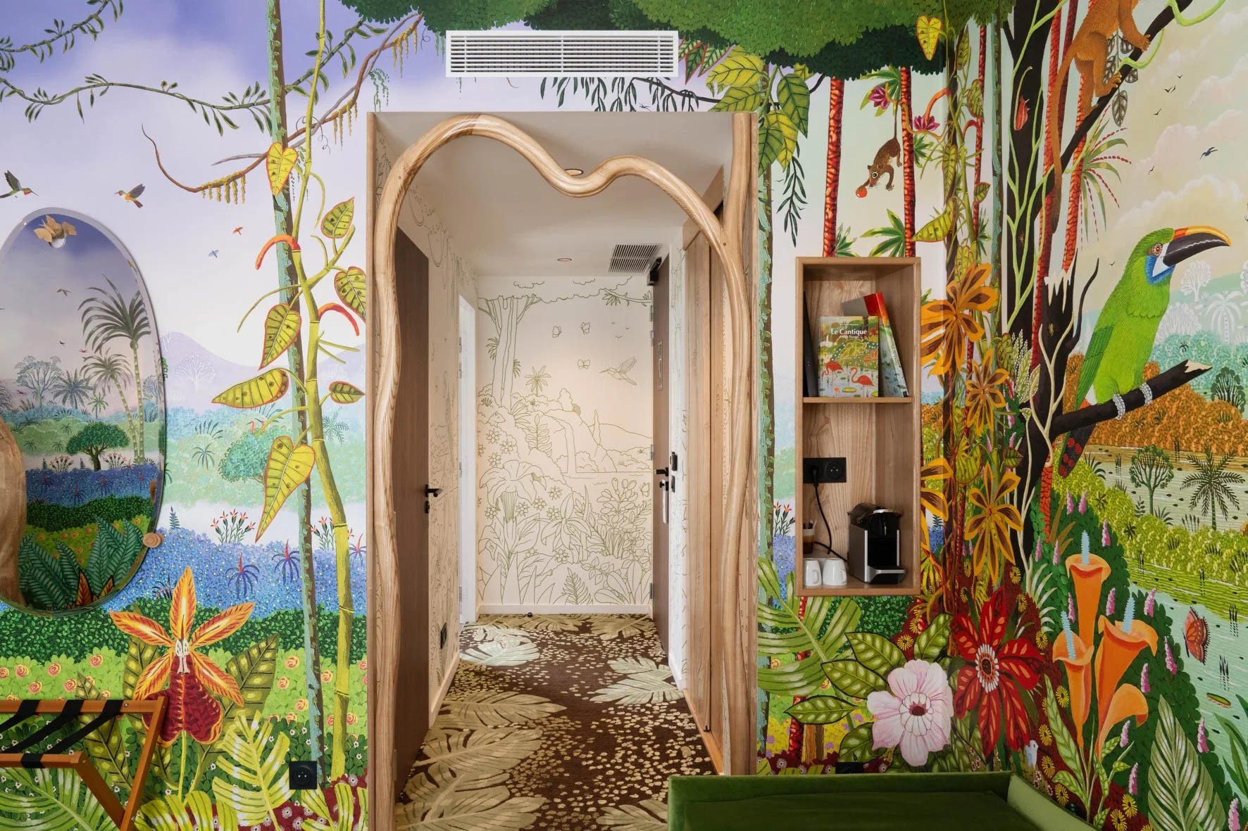 Intérieur avec mural coloré représentant une forêt tropicale avec des animaux comme un toucan et des singes, comprenant une porte en bois et une étagère contenant un livre, une cafetière et des tasses.