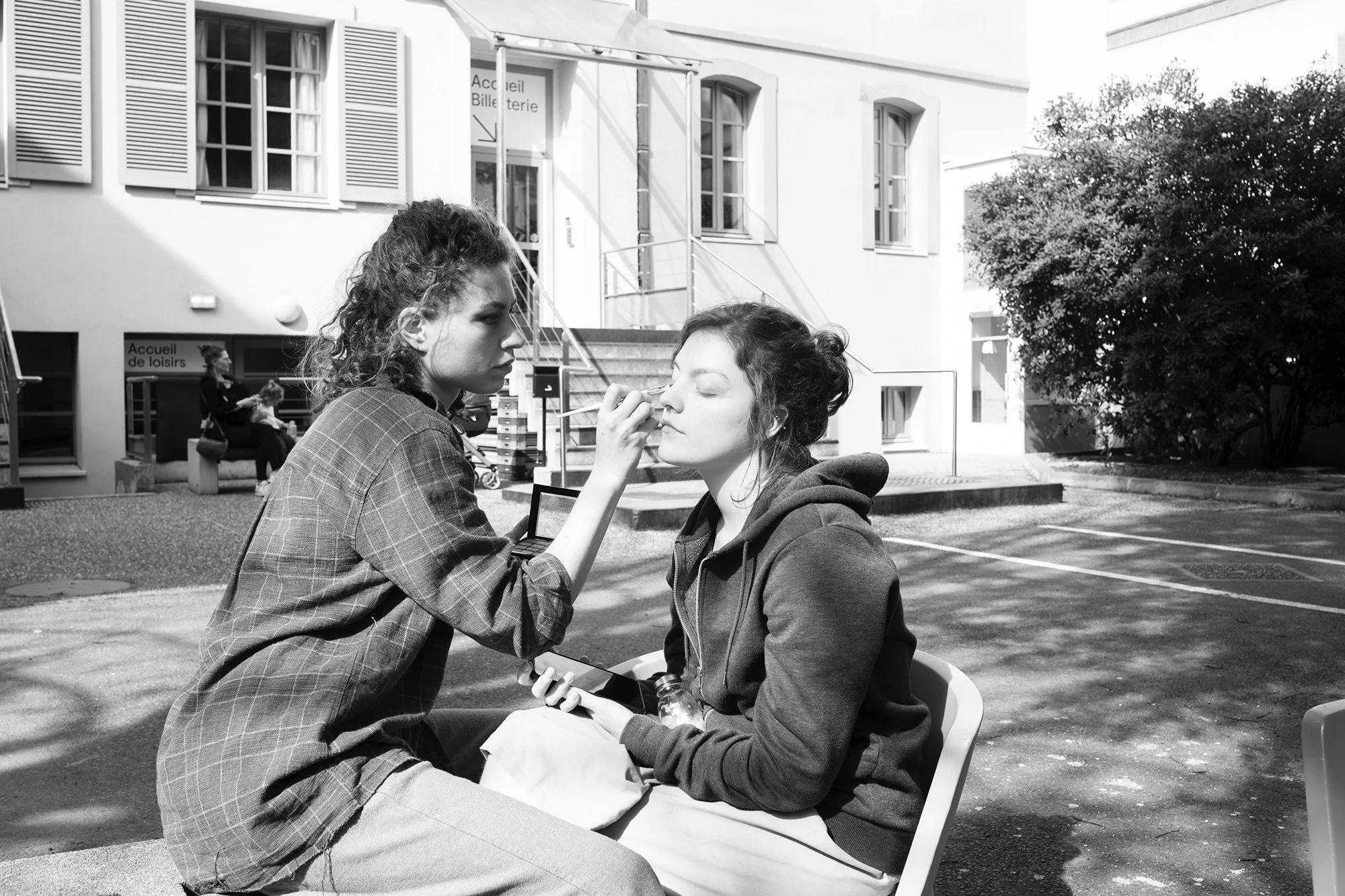 Deux jeunes femmes, une avec des cheveux bouclés foncés et l'autre avec des cheveux longs, assises à l'extérieur d'un bâtiment en train d'appliquer du maquillage à l'autre.