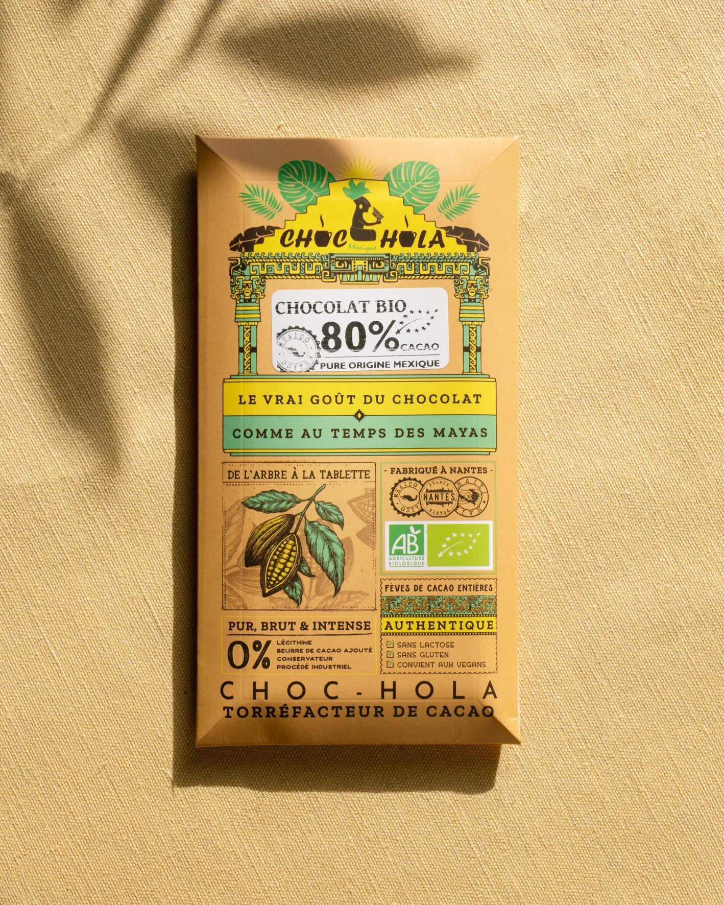 Une tablette de chocolat biologique de couleur beige avec des détails colorés, posée sur un tissu beige. La tablette comporte des textes en français, des logos biologiques, une illustration de cacaoyer, et diverses mentions sur l'origine et la composition du chocolat.