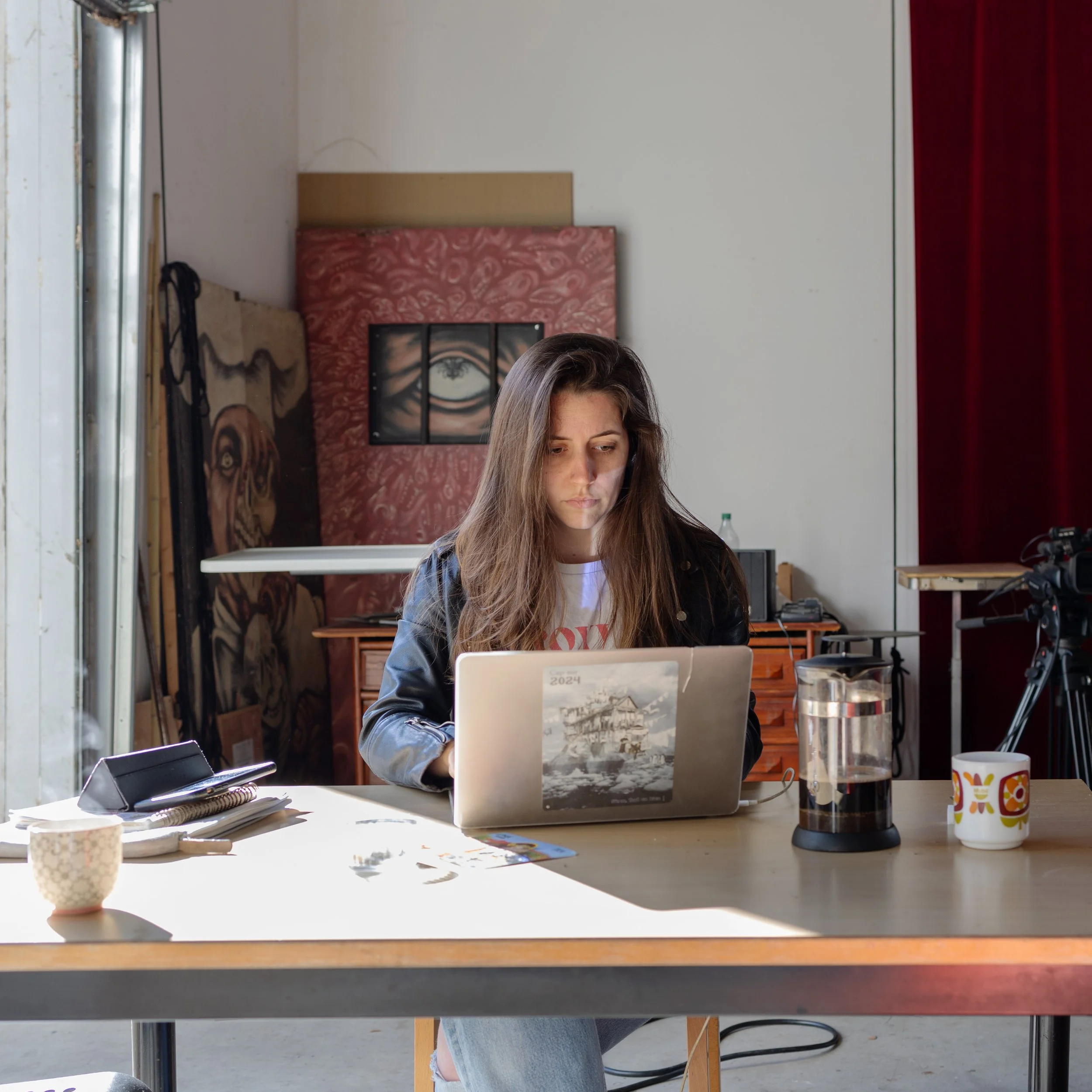 Une femme avec des cheveux bruns longs assise à une table, travaillant sur un ordinateur portable blanc dans un espace intérieur lumineux. La table a des objets divers dont un carnet, un téléphone, une tasse à café, et un thermos. En arrière-plan, un