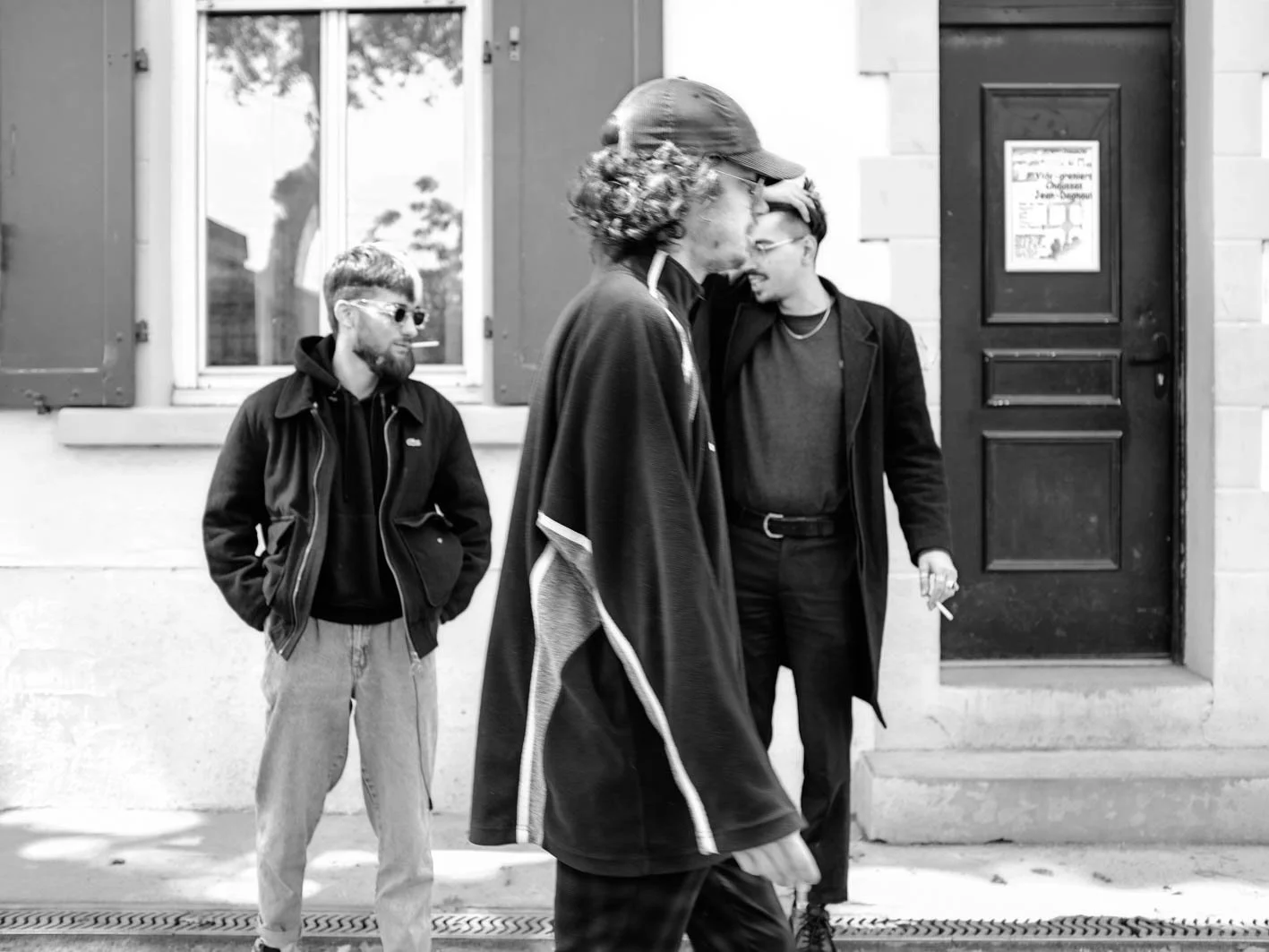Trois hommes debout sur le trottoir en face d'un bâtiment. Deux discutent et une regarde ailleurs, tous portant des vêtements décontractés.