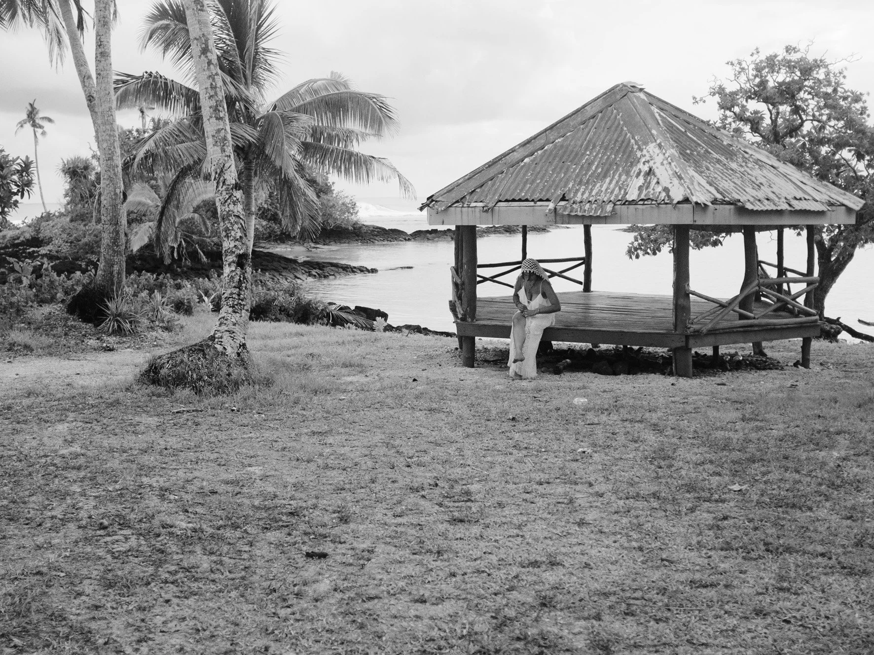 Une scène en noir et blanc d'une plage avec palmier et un gazebo en bois. Une personne est assise sur la banquette du gazebo, portant un chapeau et semblant écrire ou lire.