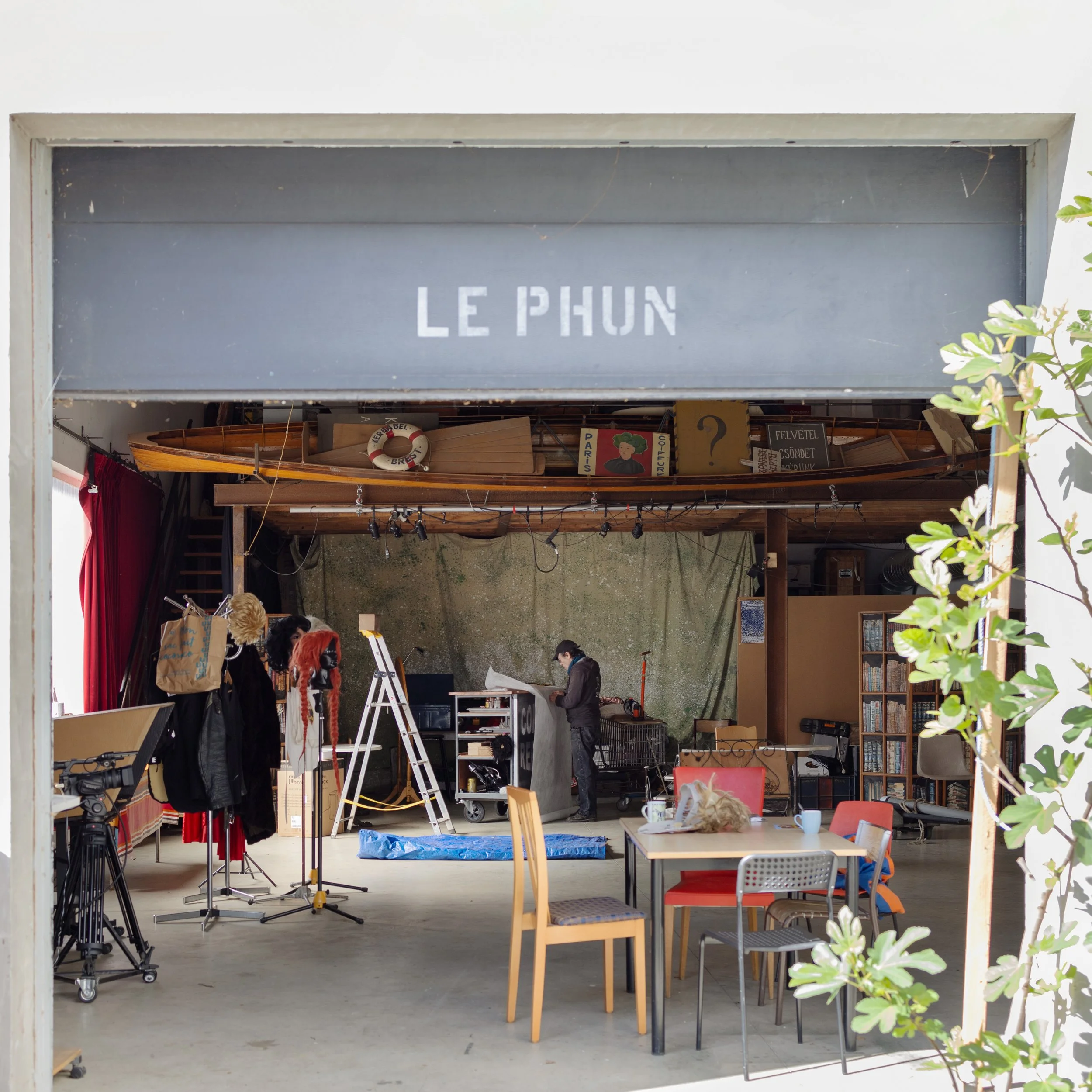 Intérieur d'une boutique ou d'un atelier avec une enseigne 'Le Phun' au-dessus, contenant un bateau en bois et diverses fournitures, des personnes portant des masques, et un décor organisé avec des meubles, des livres et un fond en béton.