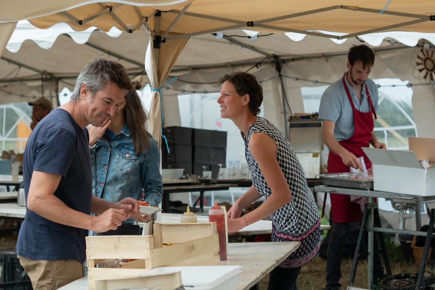 Groupe de personnes dans un stand de nourriture lors d'un événement en plein air, avec un homme en train de servir un plat et une femme souriante en train de prendre la commande.