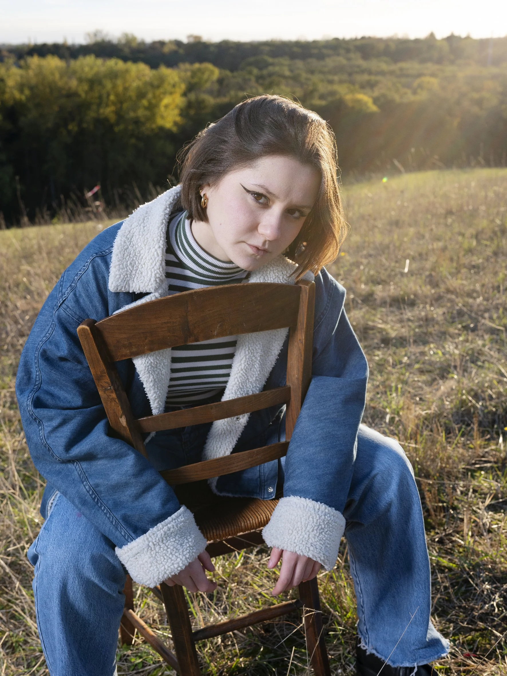 Jeune femme aux cheveux courts, portant un jean et une veste en denim avec doublure blanche, assise sur une chaise en plein air dans un champ avec des arbres au loin, sous la lumière du soleil couchant.