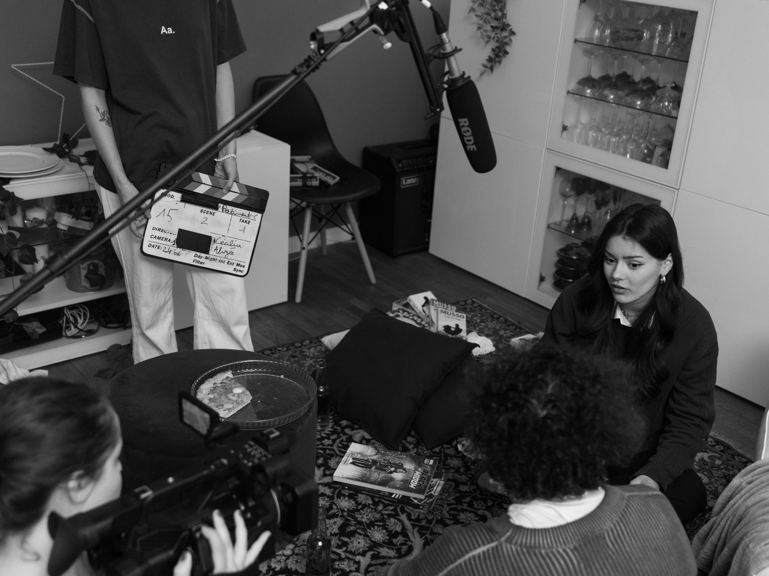 Une scène de tournage avec des personnes assises autour d'une table basse, filmée par une caméra et un micro, dans une pièce avec des étagères contenant des verres et des objets décoratifs.