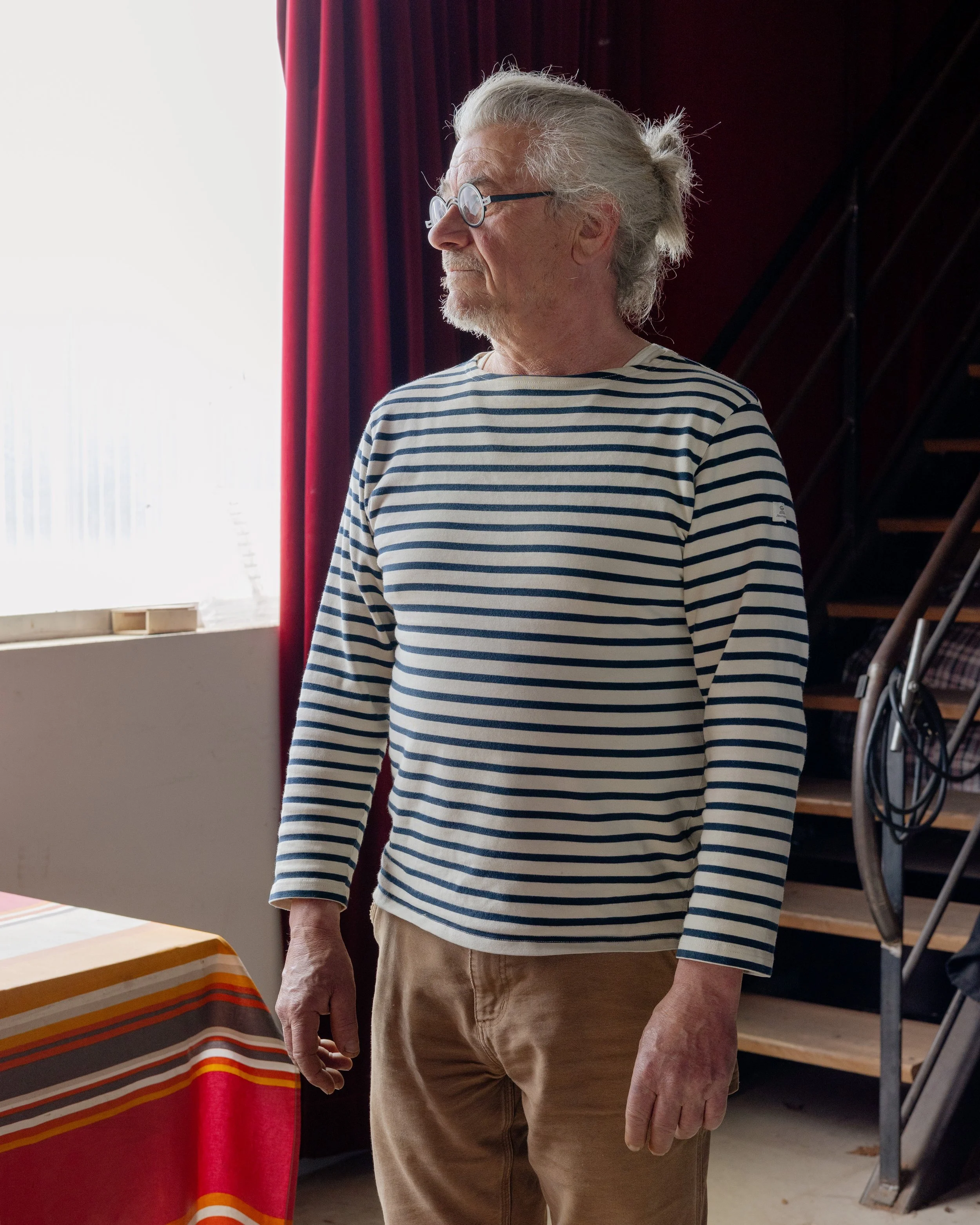 Un homme âgé avec des cheveux gris, portant des lunettes rondes et une chemise à rayures bleues et blanches, se tenant à côté d'une table à dîner avec un drap rayé orange, rouge et blanc, dans une pièce éclairée par la lumière naturelle. En arrière-p