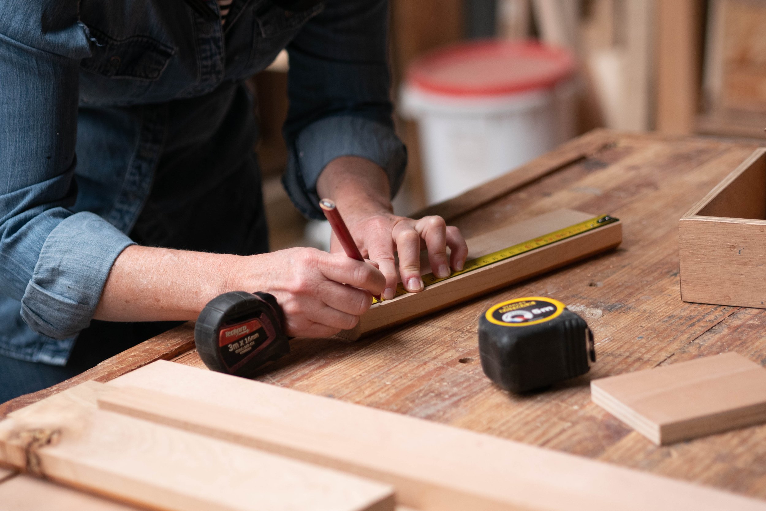 Une personne travaille sur un projet de menuiserie ou de bricolage, mesurant une pièce de bois avec un ruban à mesurer sur un établi en bois.