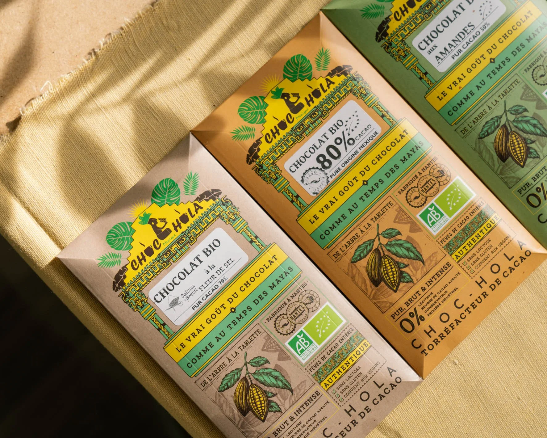 Deux paquets de chocolat bio avec des emballages colorés, l'un en beige clair et l'autre en vert, avec des illustrations de cabosse de cacao et des textes en français.