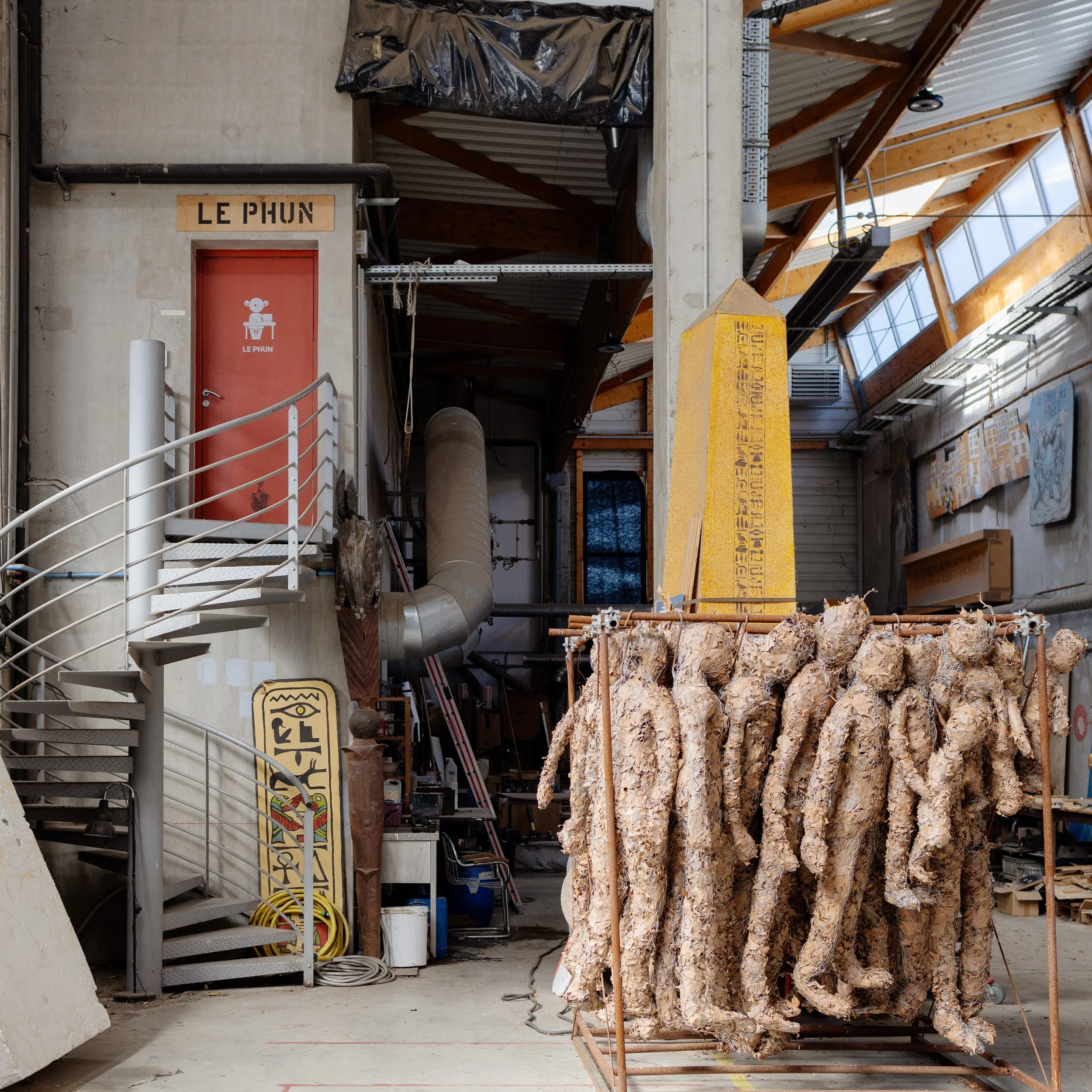 Sensation forte, un costume de pharaon en papier mâché dans un atelier d'art, entouré d'outils et de matériaux de construction, avec un escalier métallisé à gauche et un mur en béton.