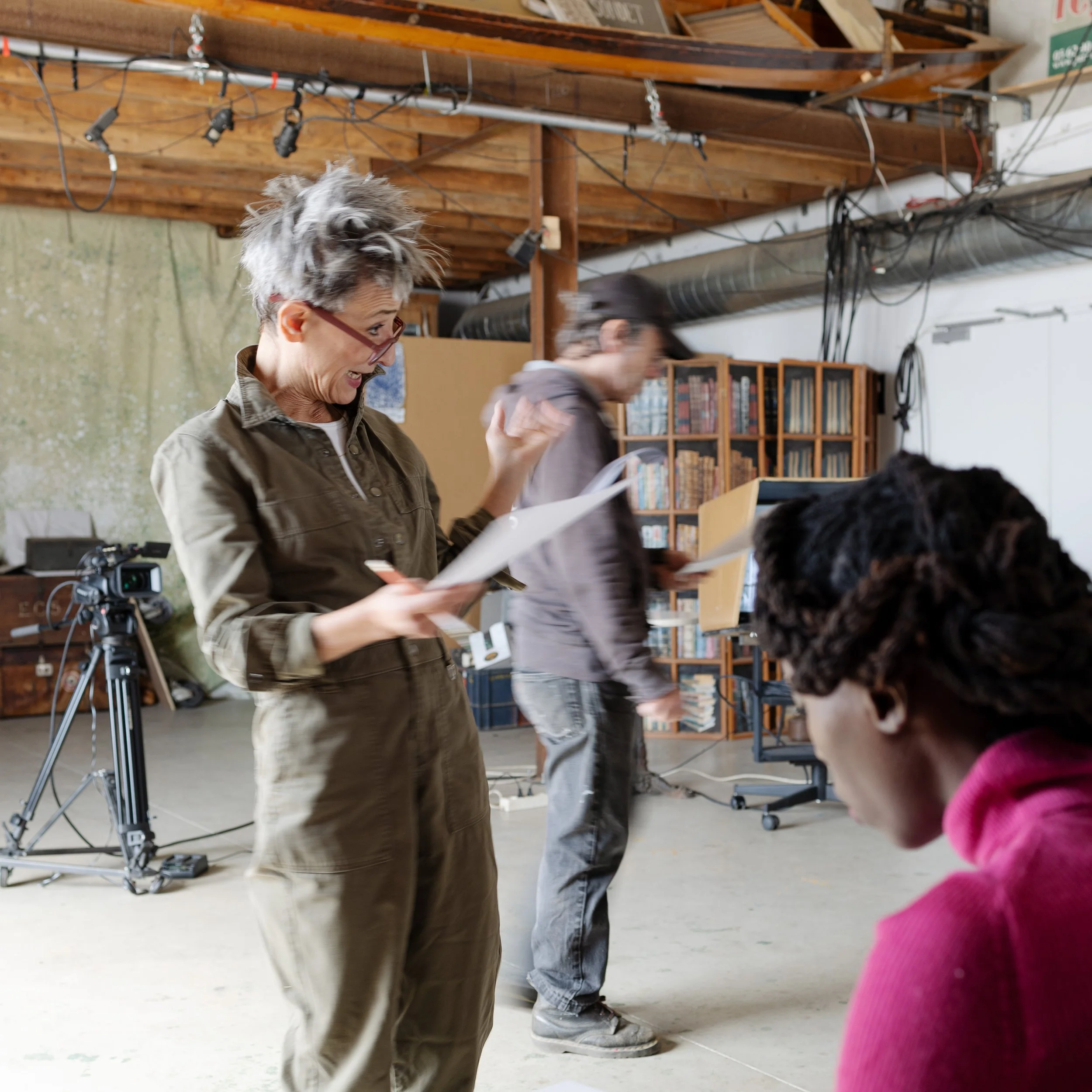 Une femme souriante avec des lunettes, portant une veste kaki, est dans un espace intérieur avec des étagères remplies de livres. Elle tient une feuille ou un document. Il y a un chien au premier plan, et quelques personnes en arrière-plan. L'espace 