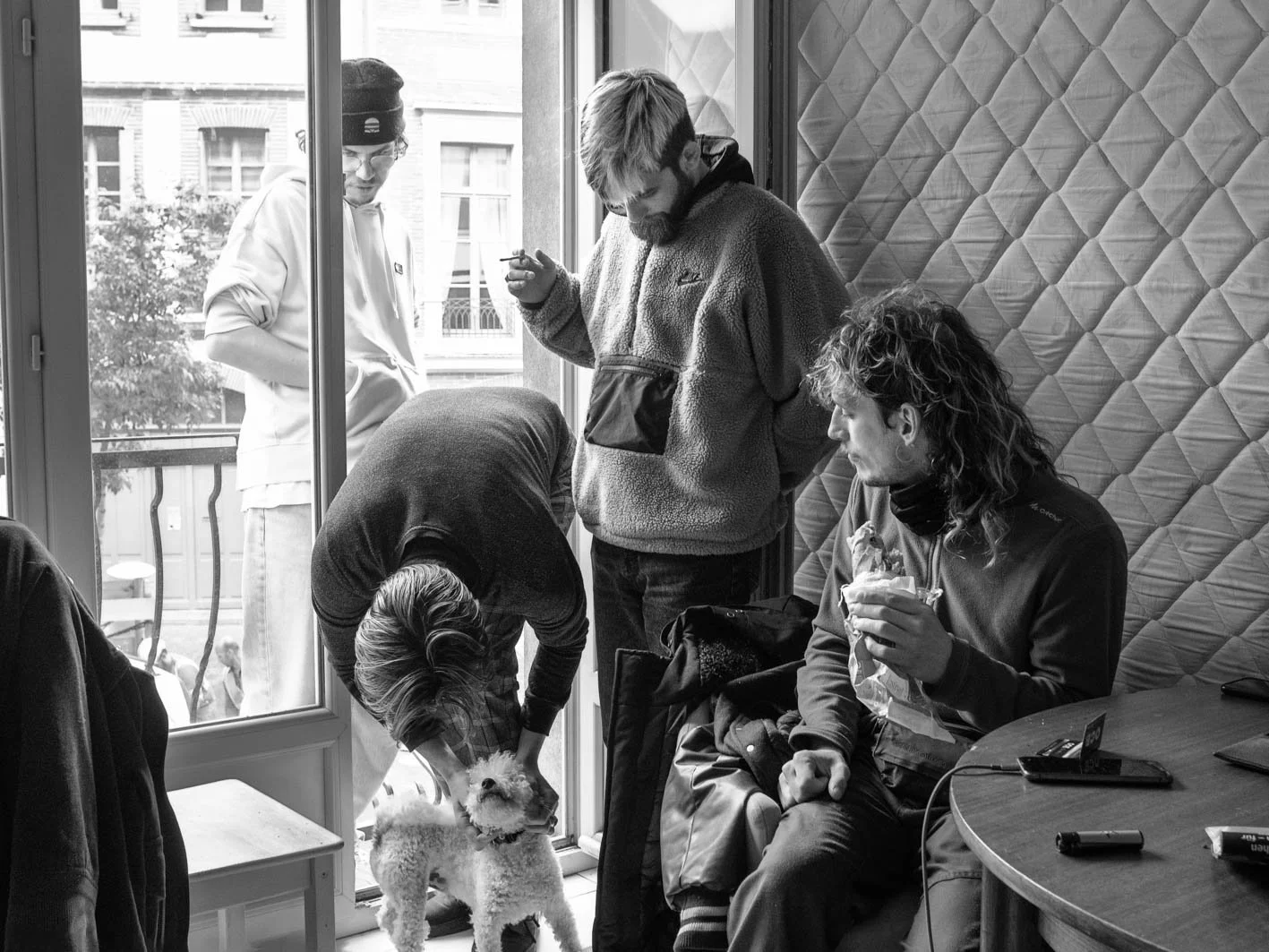 Groupe de personnes à l'intérieur d'un café ou d'une cuisine, avec un chien, près d'une fenêtre, en noir et blanc.