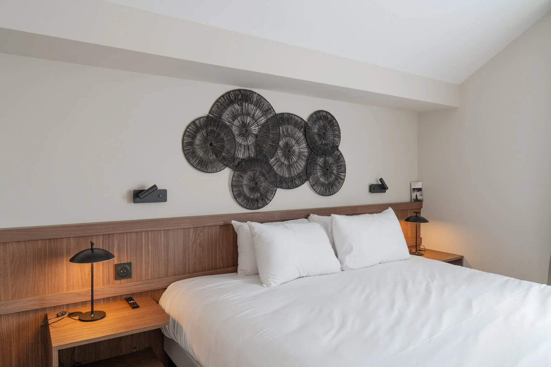 Chambre d'hôtel avec lit double, décoration minimaliste, mur blanc, décoration en métal noir au-dessus du lit, lampes de nuit modernes, table de chevet en bois.